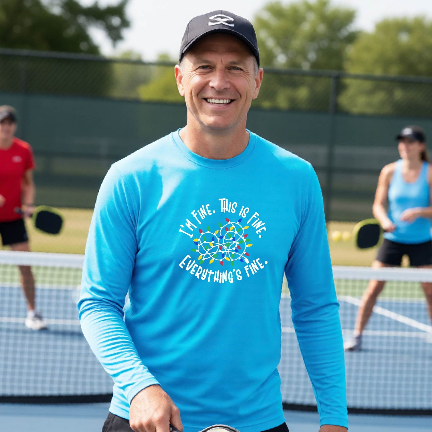 A man smiling on an pickleball court wearing a bright blue t-shirt with a colorful graphic of tangled Christmas lights and the text 'I'M FINE. THIS IS FINE. EVERYTHING'S FINE.'