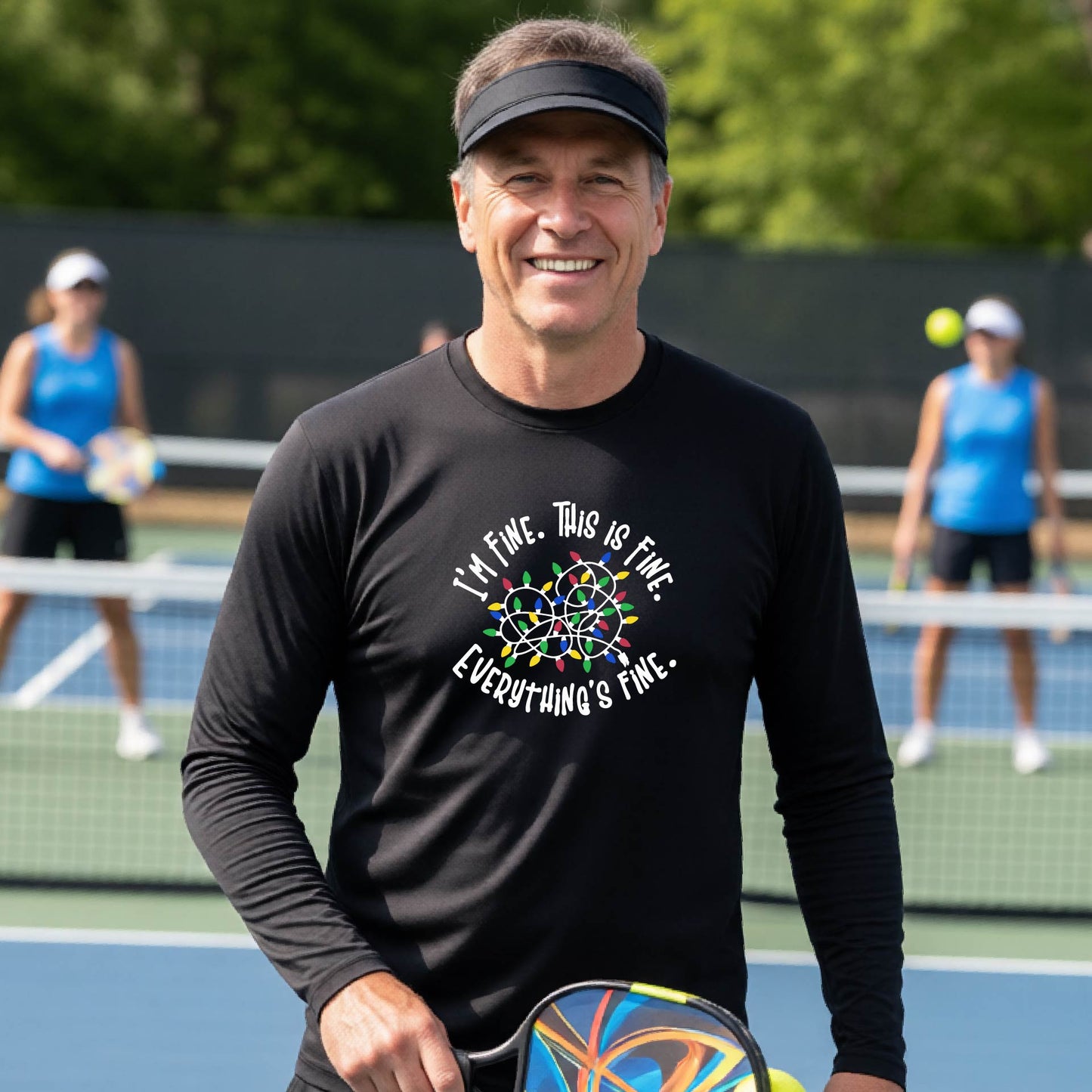 A man smiling on an pickleball court wearing a black long-sleeve t-shirt with a colorful graphic of tangled Christmas lights and the text 'I'M FINE. THIS IS FINE. EVERYTHING'S FINE.'