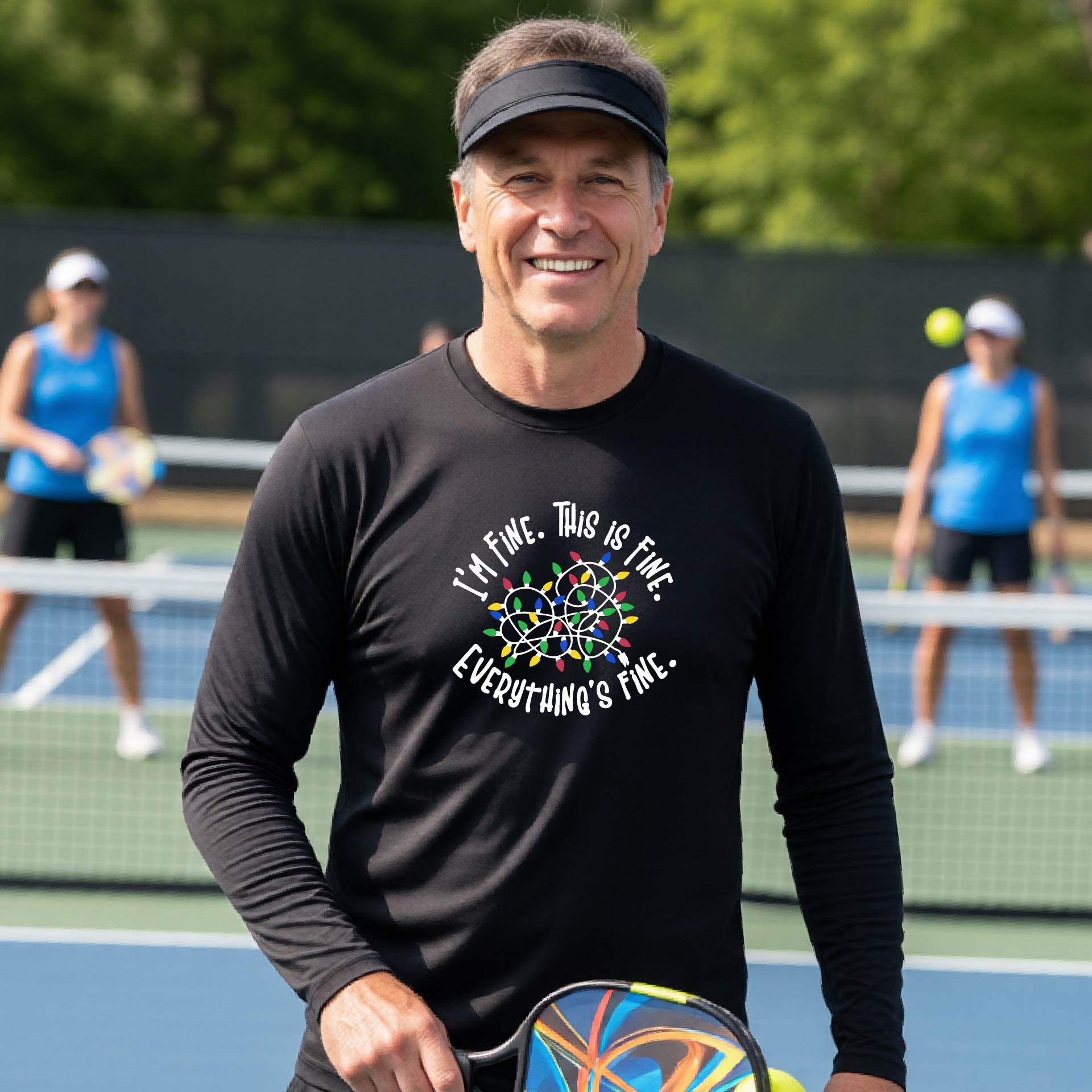 A man smiling on an pickleball court wearing a black long-sleeve t-shirt with a colorful graphic of tangled Christmas lights and the text 'I'M FINE. THIS IS FINE. EVERYTHING'S FINE.'
