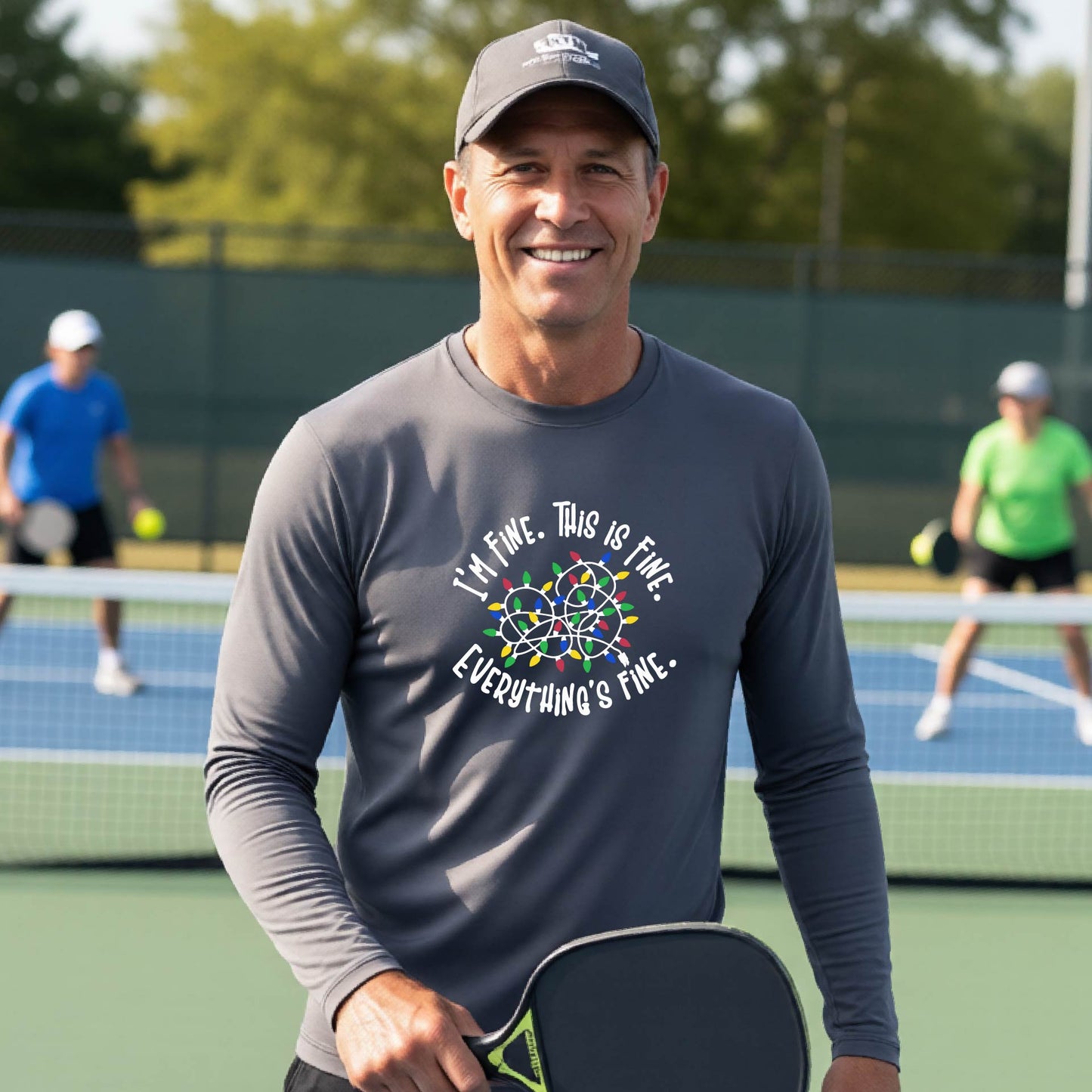 A man smiling on an pickleball court wearing a gray long-sleeve t-shirt with a colorful graphic of tangled Christmas lights and the text 'I'M FINE. THIS IS FINE. EVERYTHING'S FINE.'