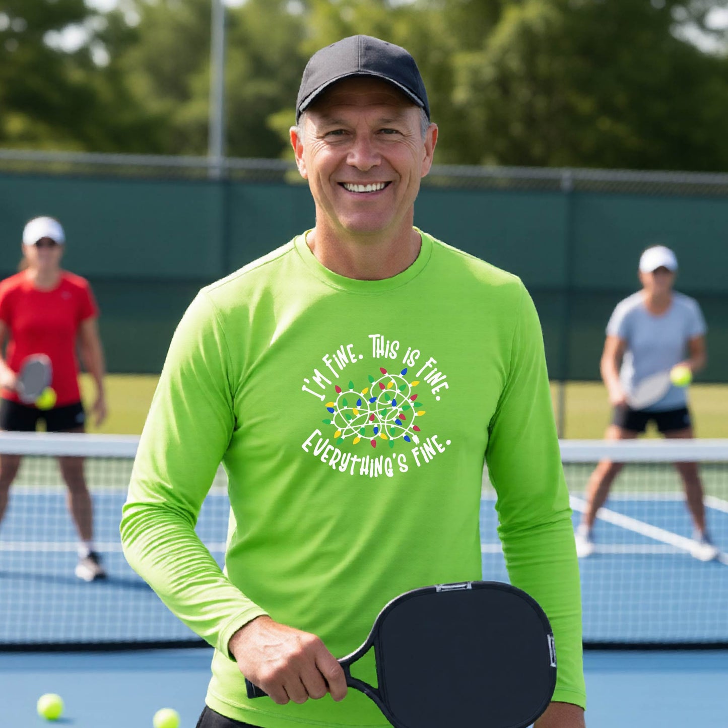 A man smiling on an pickleball court wearing a lime green long-sleeve t-shirt with a colorful graphic of tangled Christmas lights and the text 'I'M FINE. THIS IS FINE. EVERYTHING'S FINE.'