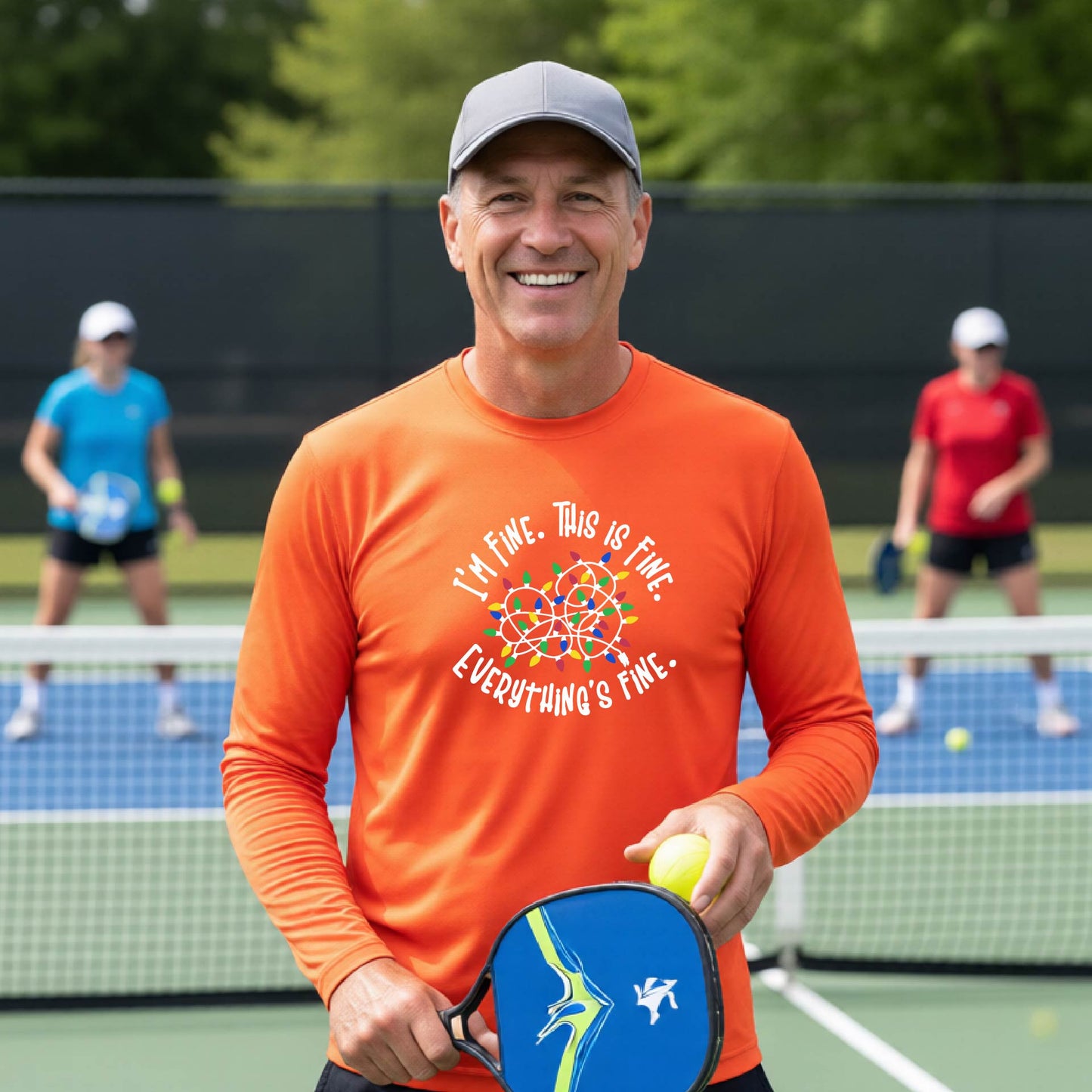 A man smiling on an pickleball court wearing an orange long-sleeve t-shirt with a colorful graphic of tangled Christmas lights and the text 'I'M FINE. THIS IS FINE. EVERYTHING'S FINE.'