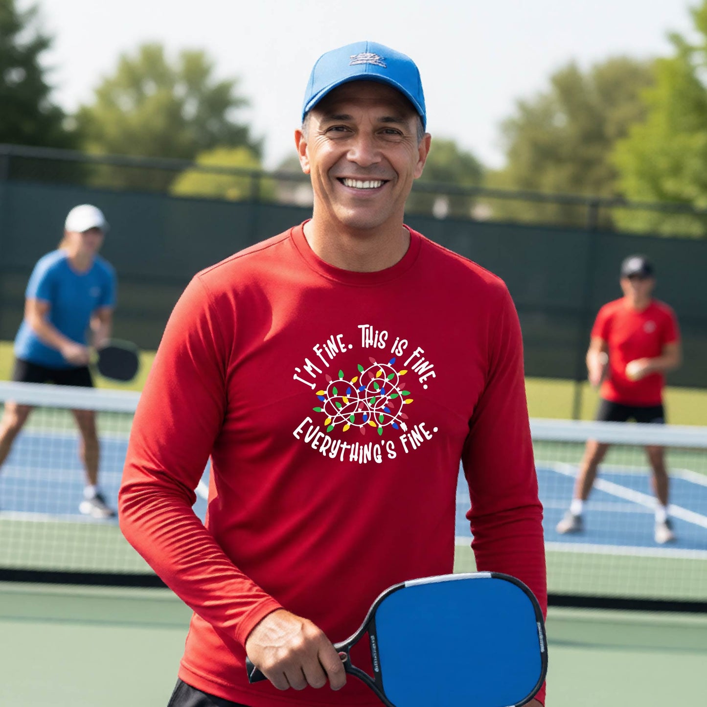 A man smiling on an pickleball court wearing a red long-sleeve t-shirt with a colorful graphic of tangled Christmas lights and the text 'I'M FINE. THIS IS FINE. EVERYTHING'S FINE.'
