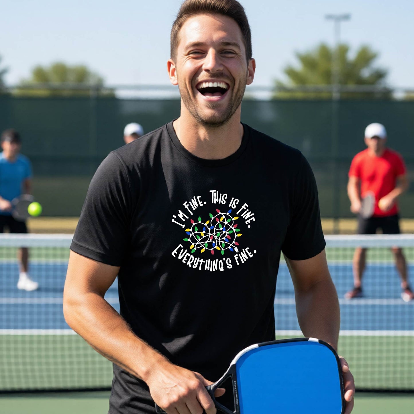 A man smiling on an pickleball court wearing a black t-shirt with a colorful graphic of tangeled Christmas lights and the text 'I'M FINE. THIS IS FINE. EVERYTHING'S FINE.'