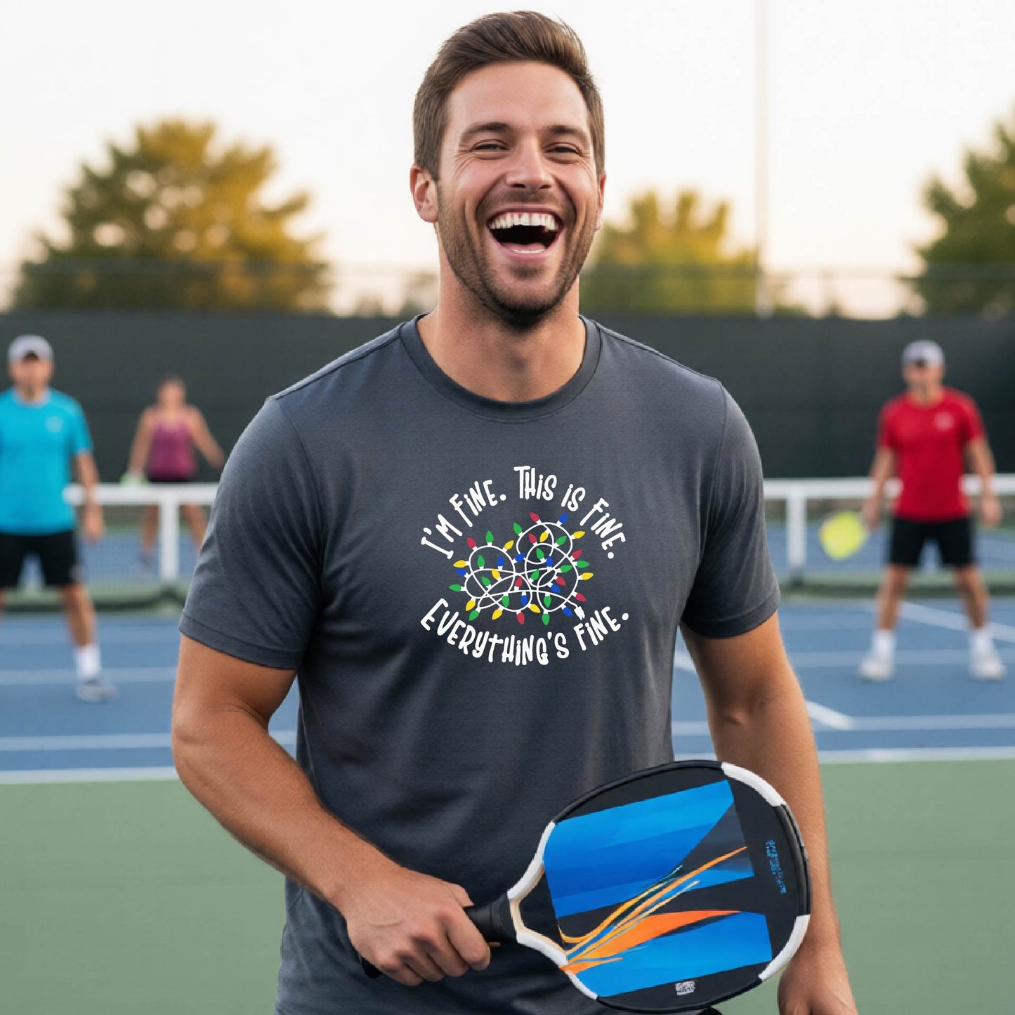 A man smiling on an pickleball court wearing a grey t-shirt with a colorful graphic of tangeled Christmas lights and the text 'I'M FINE. THIS IS FINE. EVERYTHING'S FINE.'