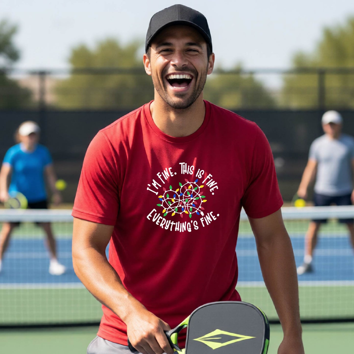 A man smiling on an pickleball court wearing a RED t-shirt with a colorful graphic of tangeled Christmas lights and the text 'I'M FINE. THIS IS FINE. EVERYTHING'S FINE.'