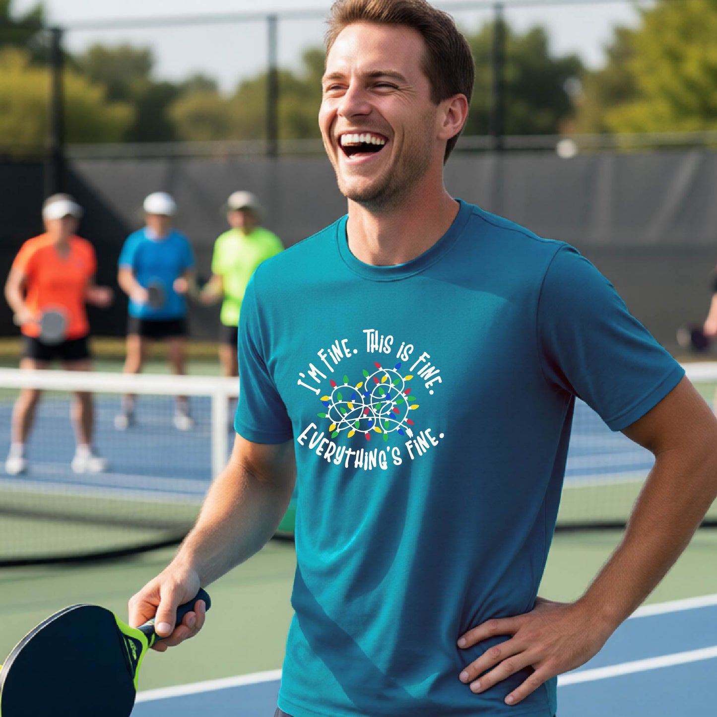 A man smiling on an pickleball court wearing a blue t-shirt with a colorful graphic of tangeled Christmas lights and the text 'I'M FINE. THIS IS FINE. EVERYTHING'S FINE.'