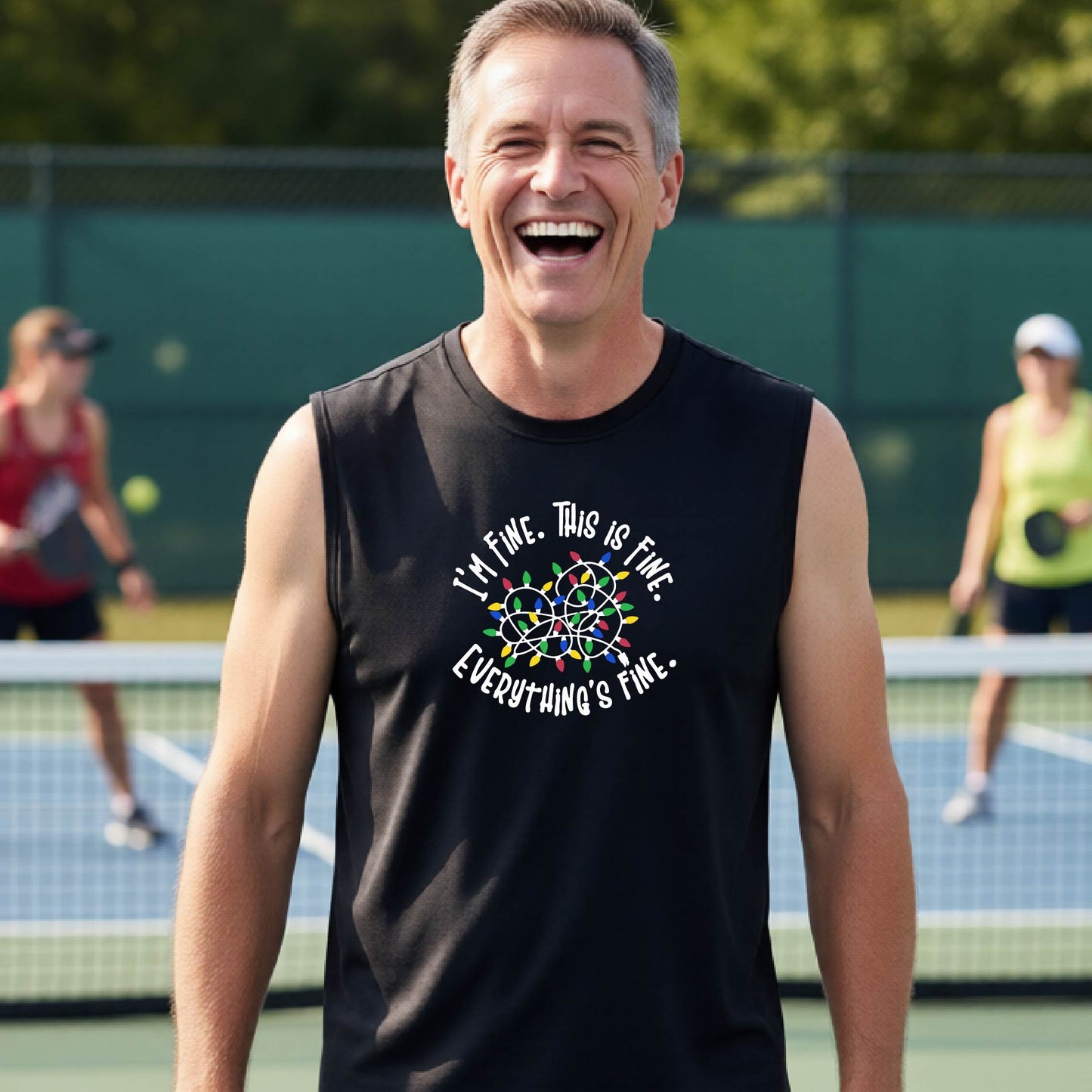 A smiling male wearing a black sleeveless t-shirt with a pickleball court in the background.  The shirt has a graphic of a tangled string of christmas lights witht the words 'I'M FINE. THIS IS FINE. EVERYTHING'S FINE.'