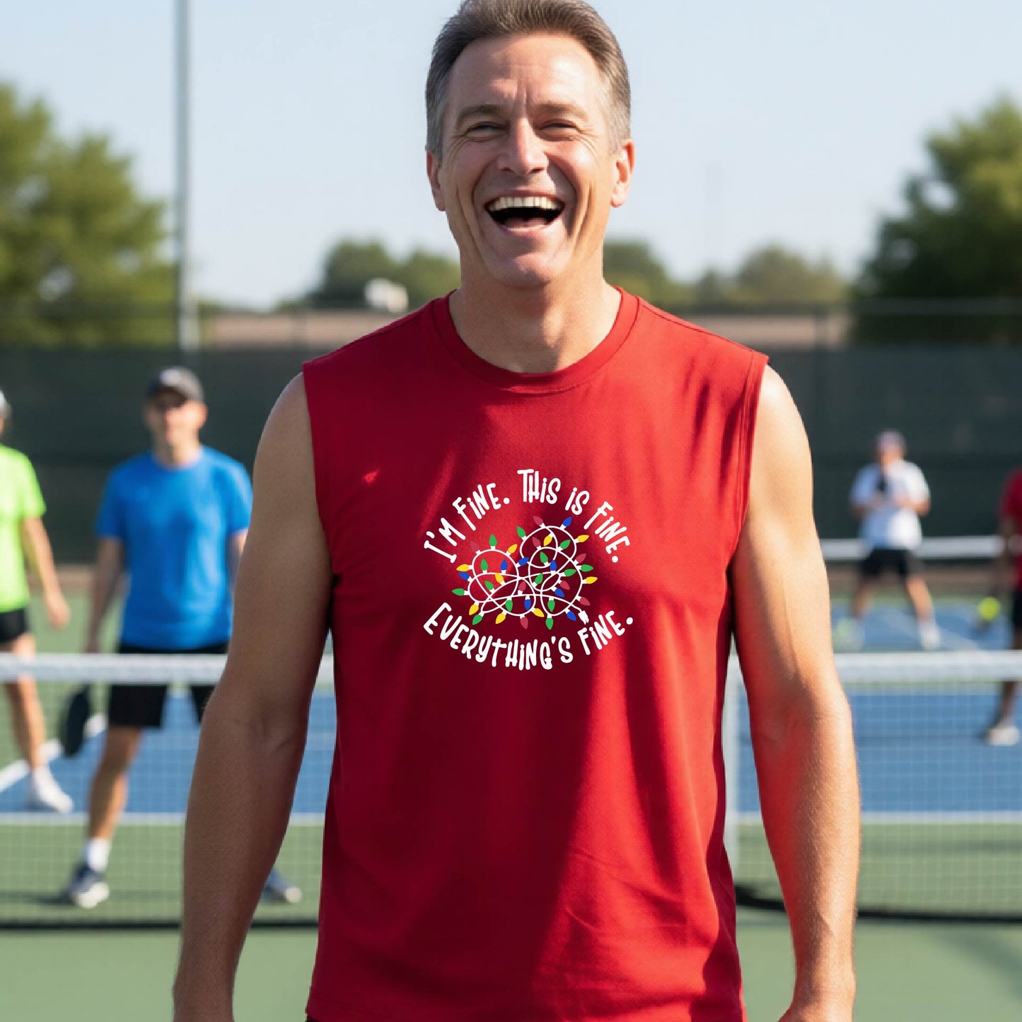 A smiling male wearing a red sleeveless t-shirt with a pickleball court in the background.  The shirt has a graphic of a tangled string of christmas lights witht the words 'I'M FINE. THIS IS FINE. EVERYTHING'S FINE.
