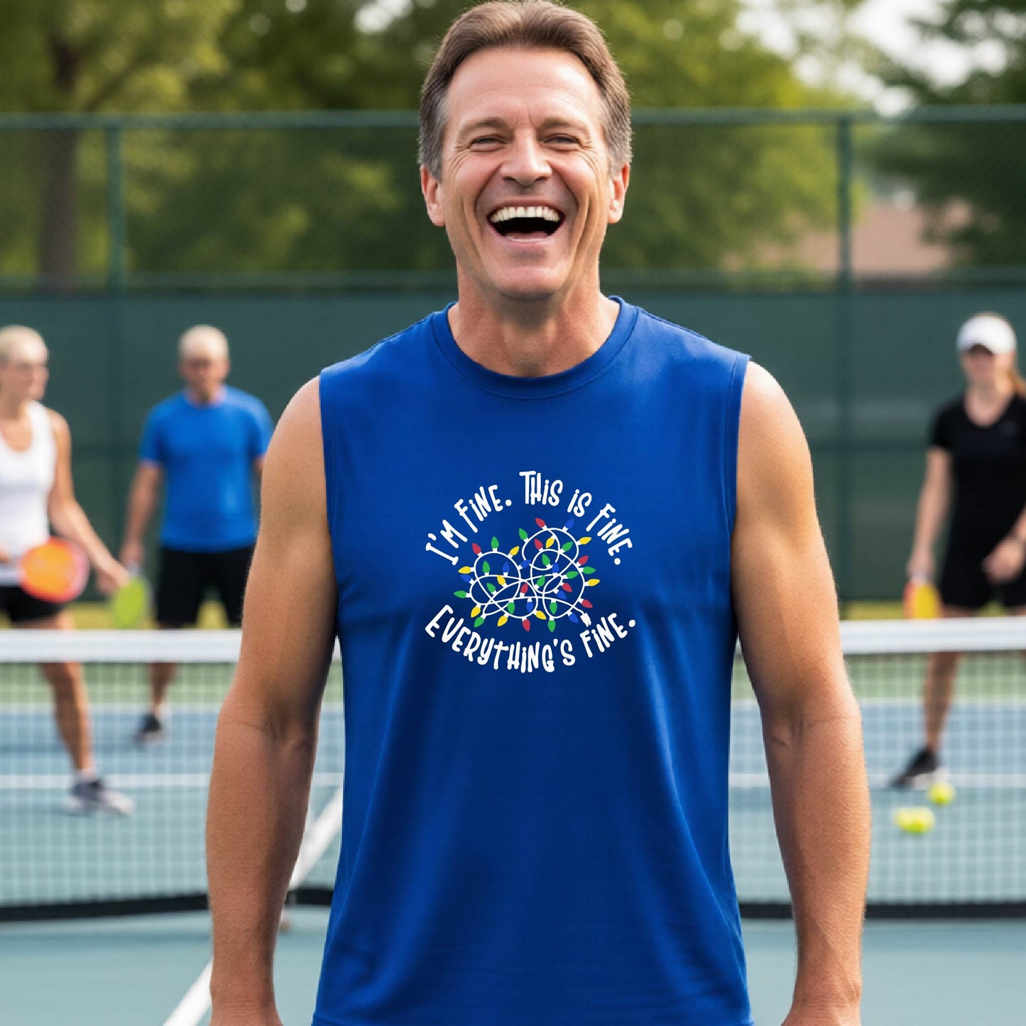 A smiling male wearing a royal blue sleeveless t-shirt with a pickleball court in the background.  The shirt has a graphic of a tangled string of christmas lights witht the words 'I'M FINE. THIS IS FINE. EVERYTHING'S FINE.