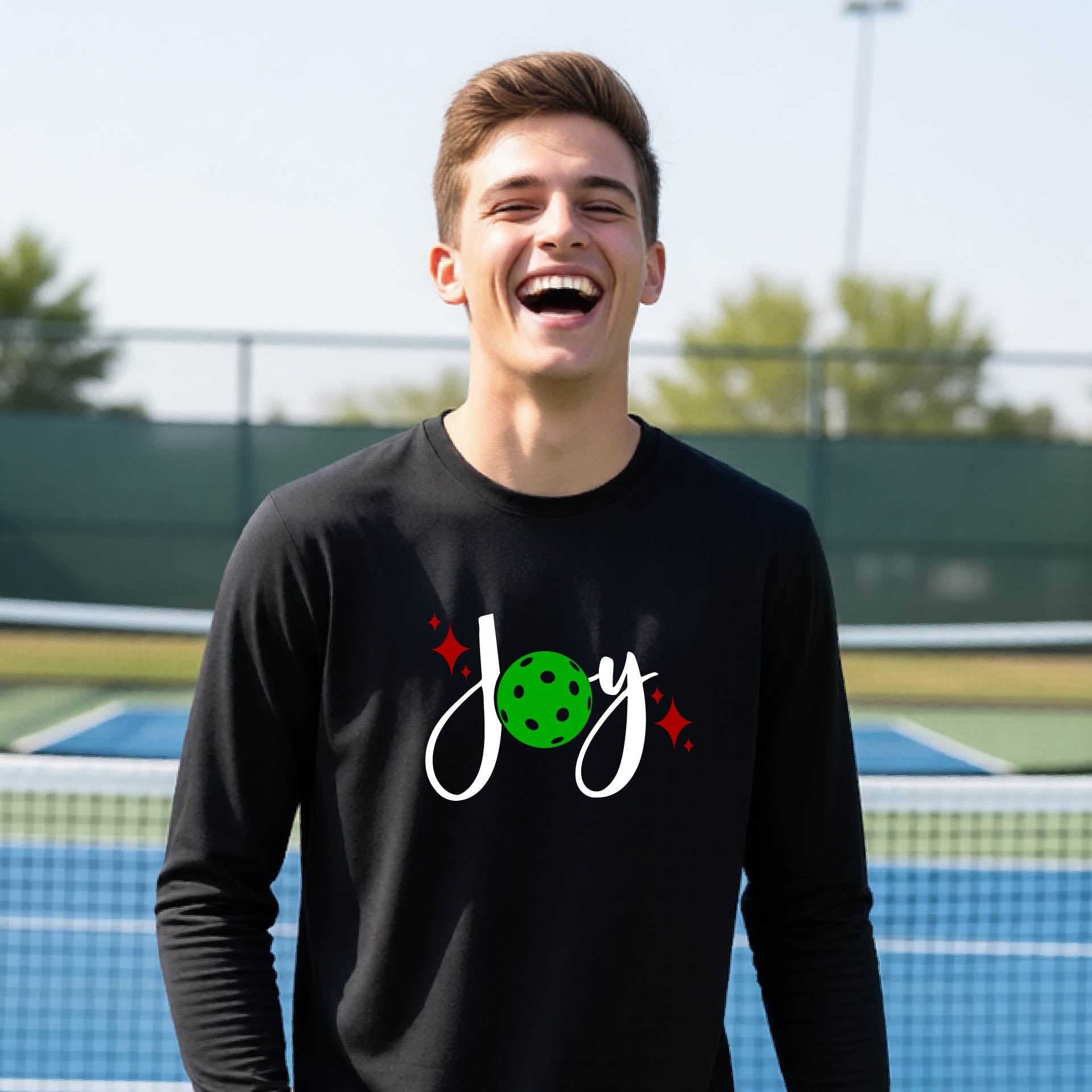 A young person laughing on an outdoor pickleball court wearing a black long-sleeve shirt with a 'joy' pickleball graphic in white and  green. 