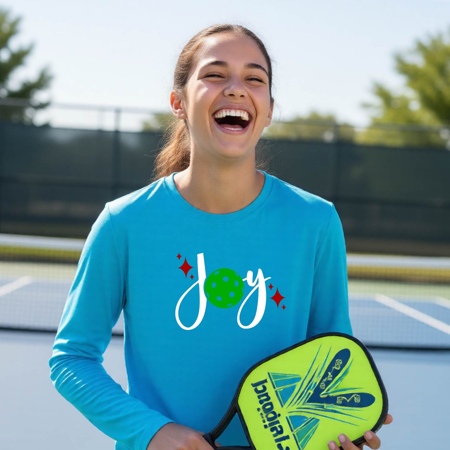 A young person laughing on an outdoor pickleball court wearing a bright blue long-sleeve shirt with a 'joy' pickleball graphic in white and  green. 