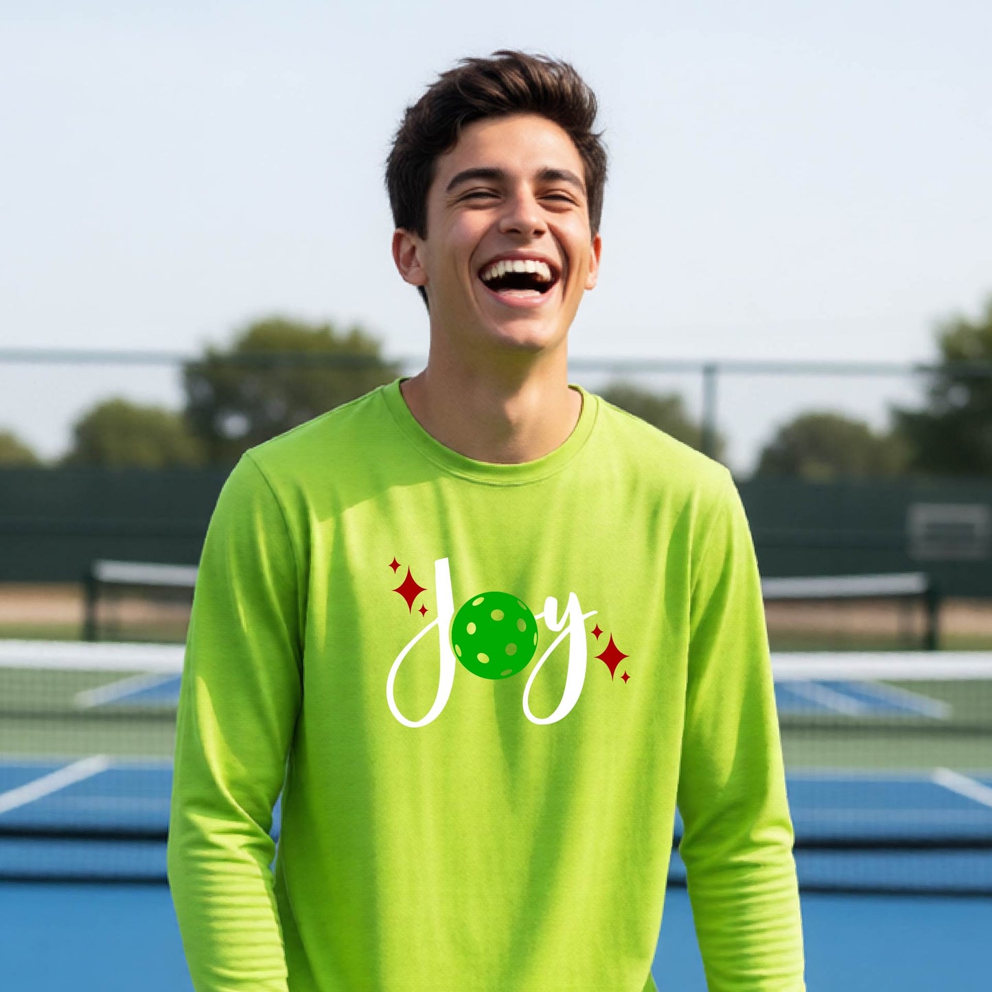 A young person laughing on an outdoor pickleball court wearing a bright lime green long-sleeve shirt with a 'joy' pickleball graphic in white and green.