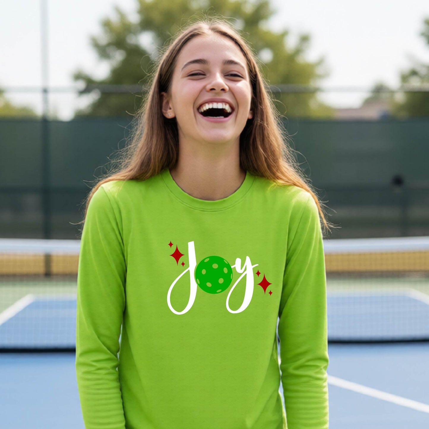 A young person laughing on an outdoor pickleball court wearing a lime green long-sleeve shirt with a 'joy' pickleball graphic in white and  green. 