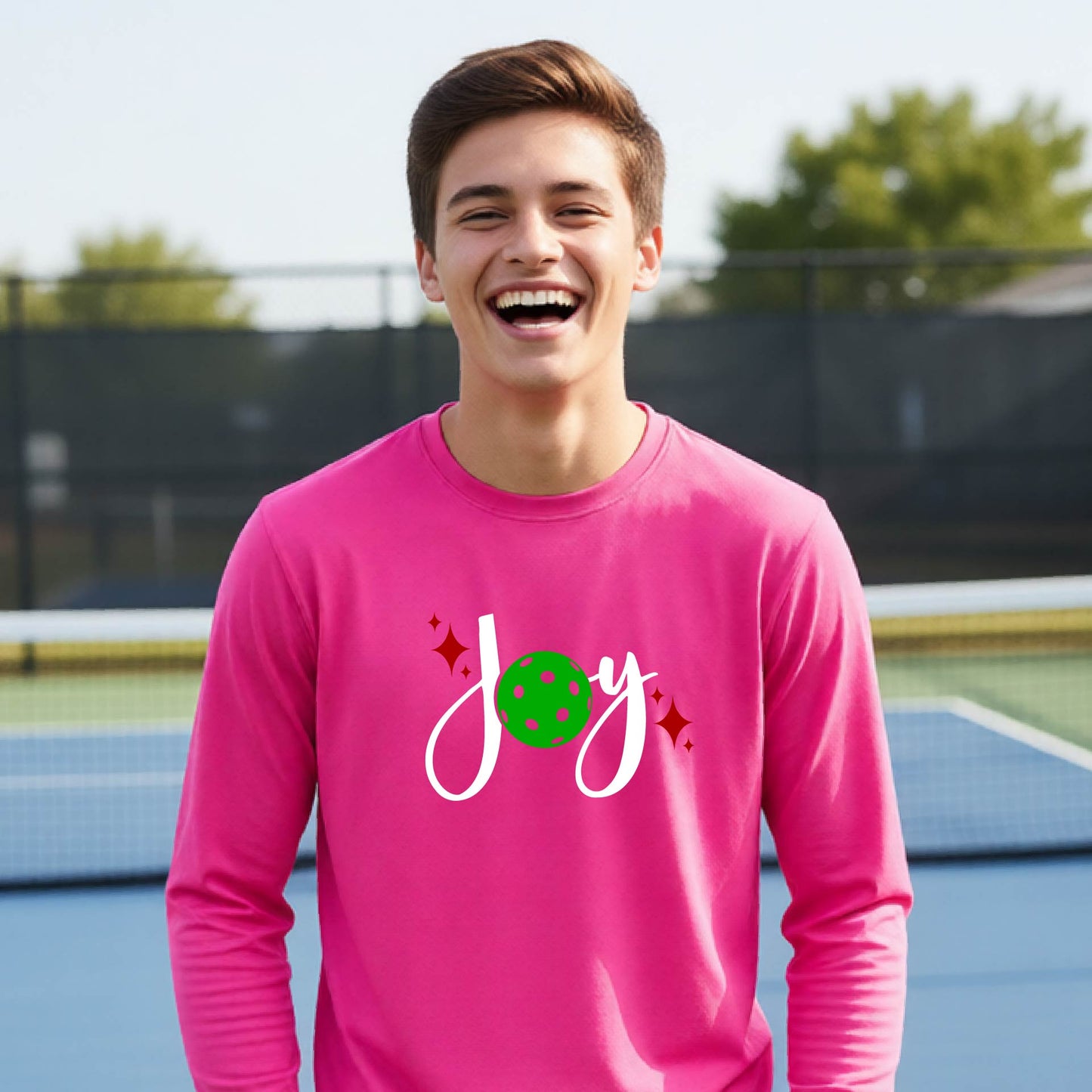 A young person laughing on an outdoor pickleball court wearing a pink long-sleeve shirt with a 'joy' pickleball graphic in white and  green. 