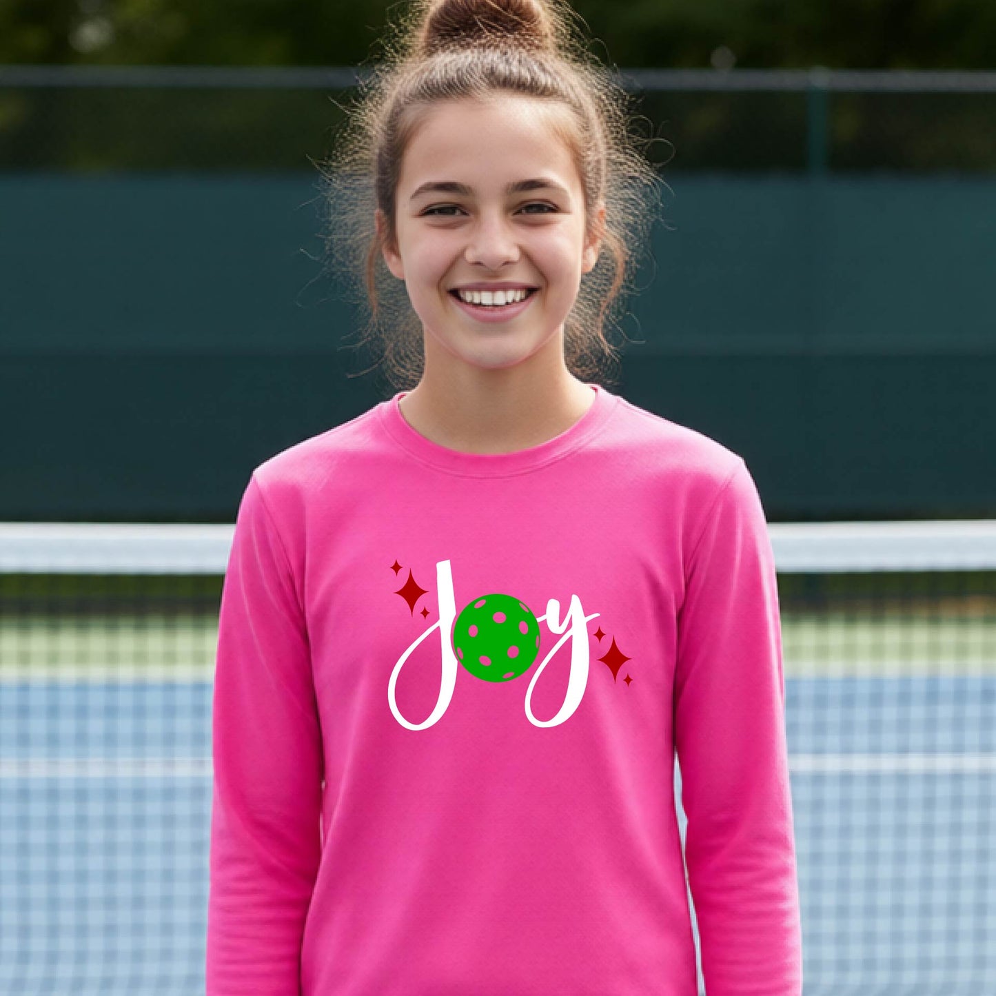 A young person laughing on an outdoor pickleball court wearing a pink long-sleeve shirt with a 'joy' pickleball graphic in white and  green. 
