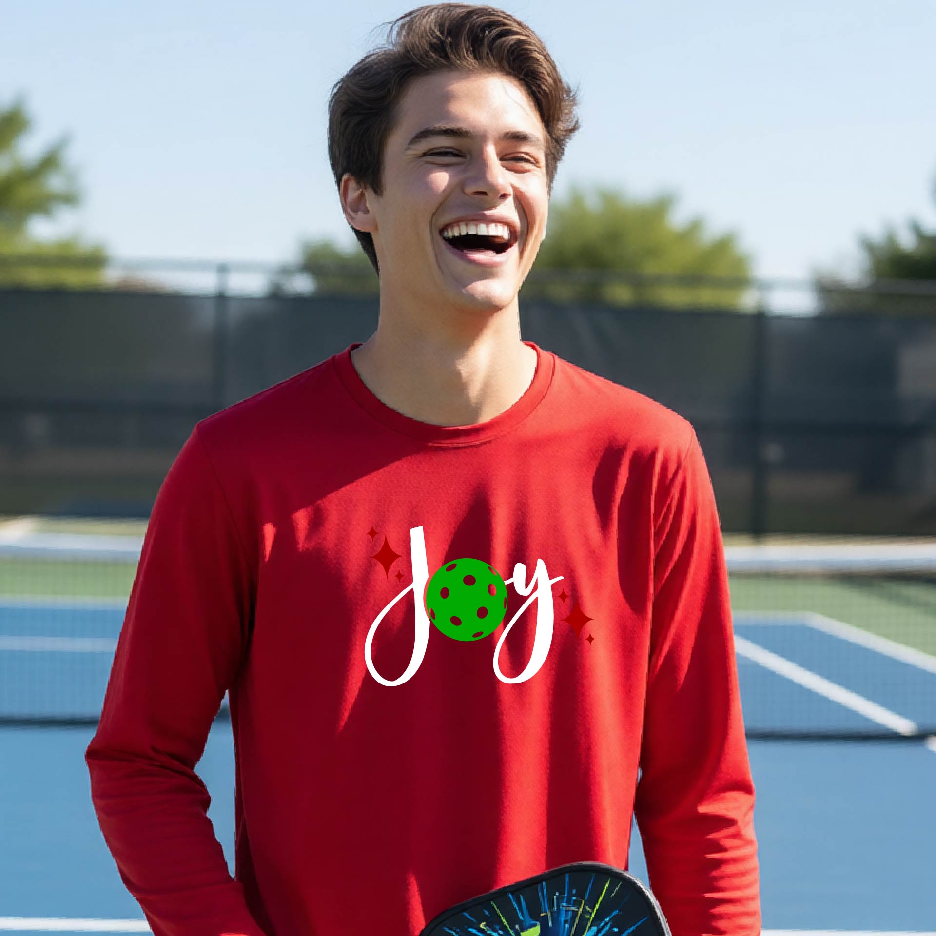A young person laughing on an outdoor pickleball court wearing a red long-sleeve shirt with a 'joy' pickleball graphic in white and  green. 