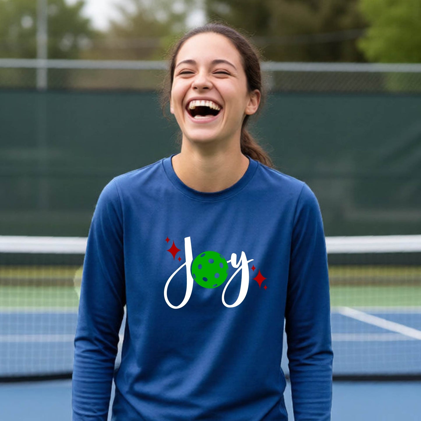 A young person laughing on an outdoor pickleball court wearing a royal blue long-sleeve shirt with a 'joy' pickleball graphic in white and  green. 