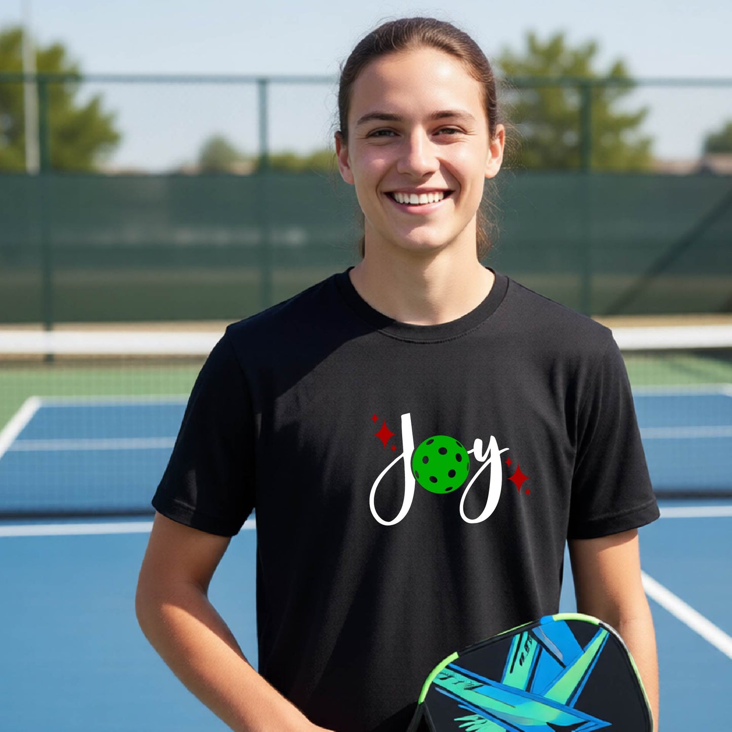 A young person laughing on an outdoor pickleball  court wearing a black t-shirt with a white 'Joy' graphic and a green pickleball  motif.