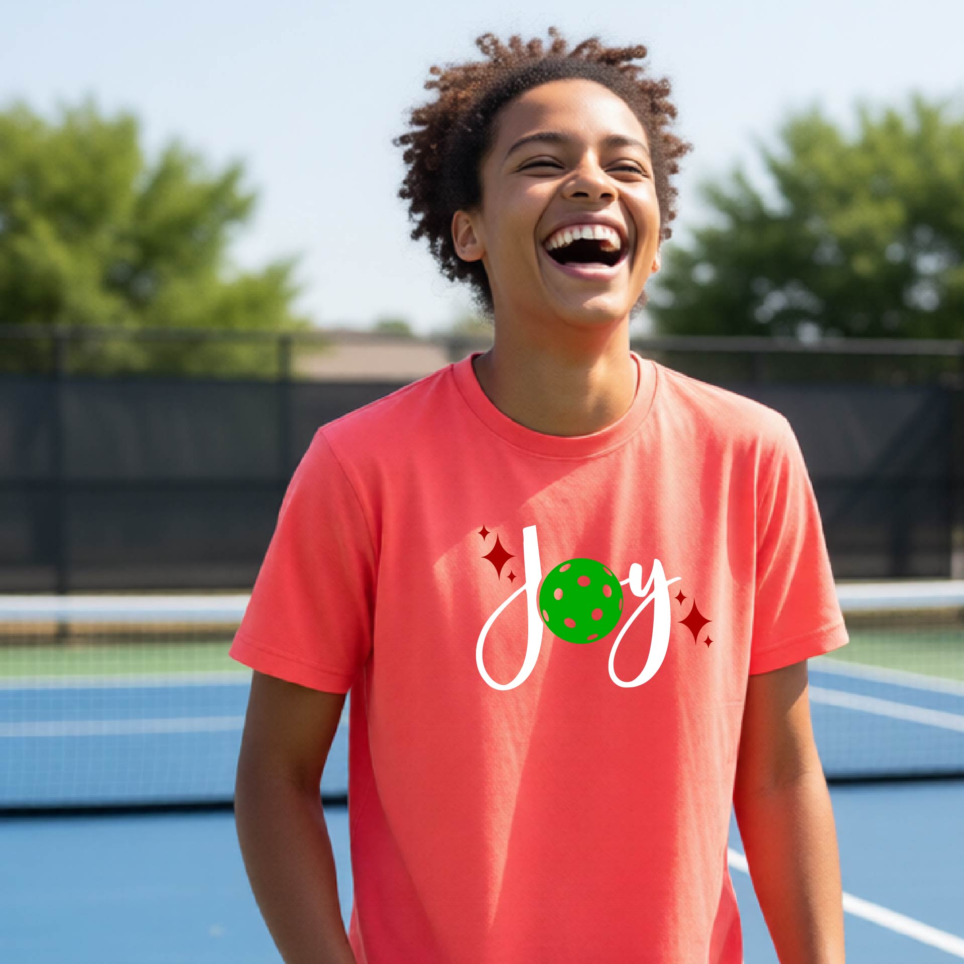 A young person laughing on an outdoor pickleball  court wearing a coral-colored t-shirt with a white 'Joy' graphic and a green pickleball  motif.