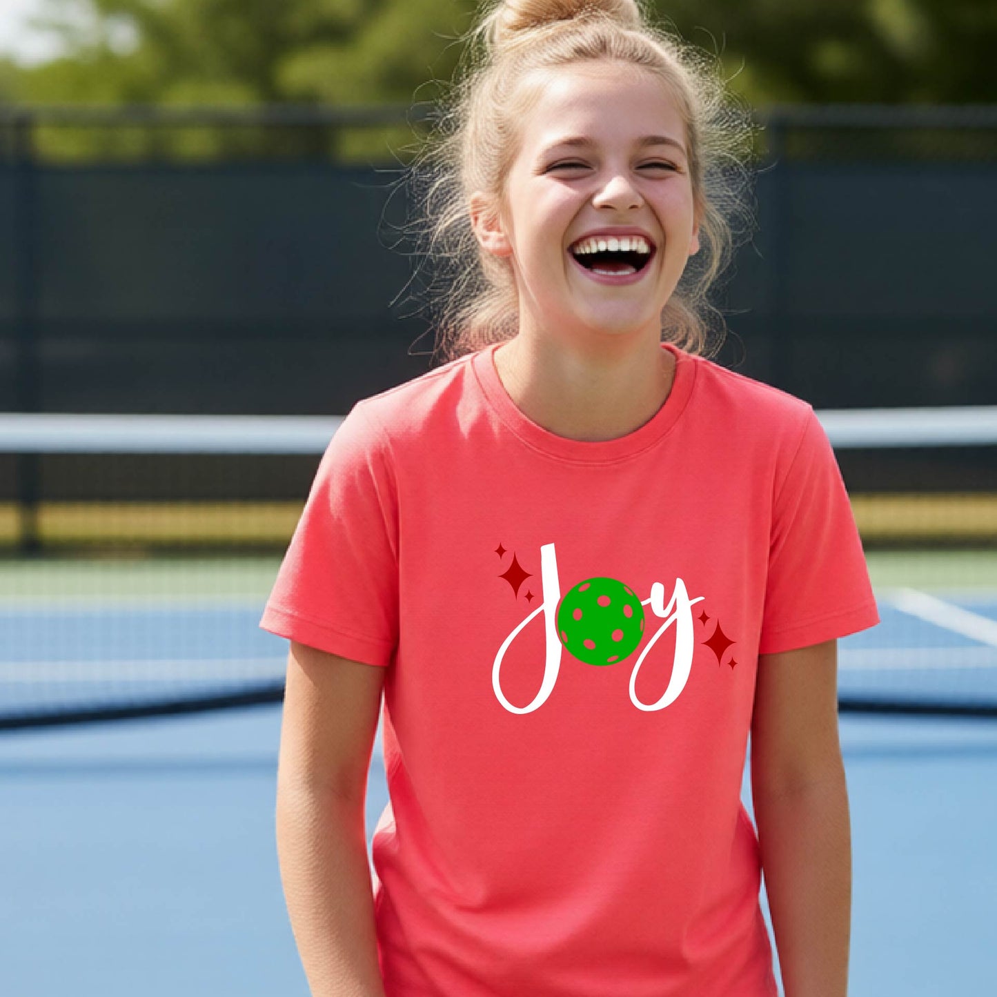 A young person laughing on an outdoor pickleball  court wearing a coral-colored t-shirt with a white 'Joy' graphic and a green pickleball  motif.