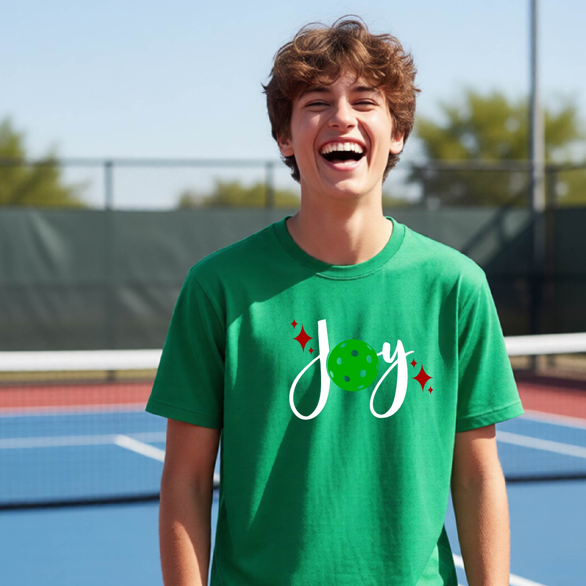A young person laughing on an outdoor pickleball  court wearing a green t-shirt with a white 'Joy' graphic and a green pickleball  motif.