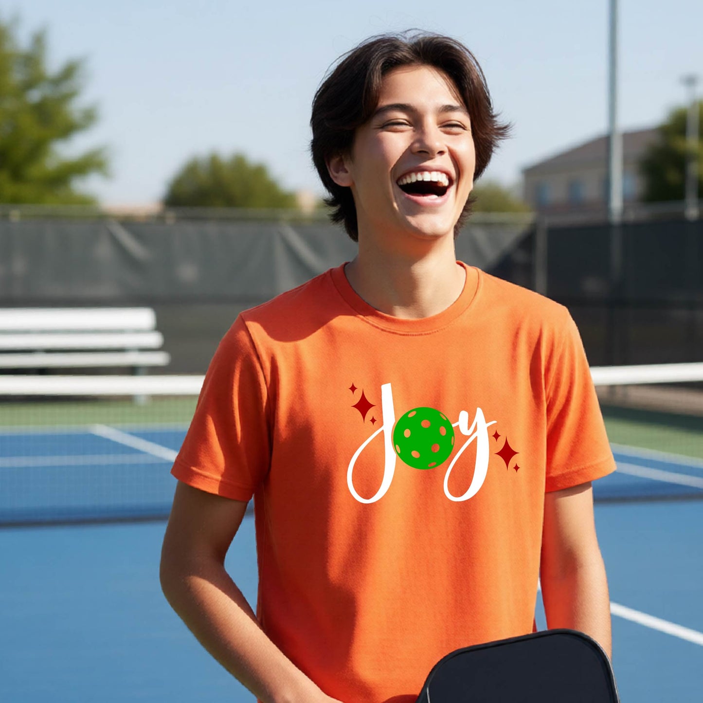 A young person laughing on an outdoor pickleball  court wearing an orange t-shirt with a white 'Joy' graphic and a green pickleball  motif.
