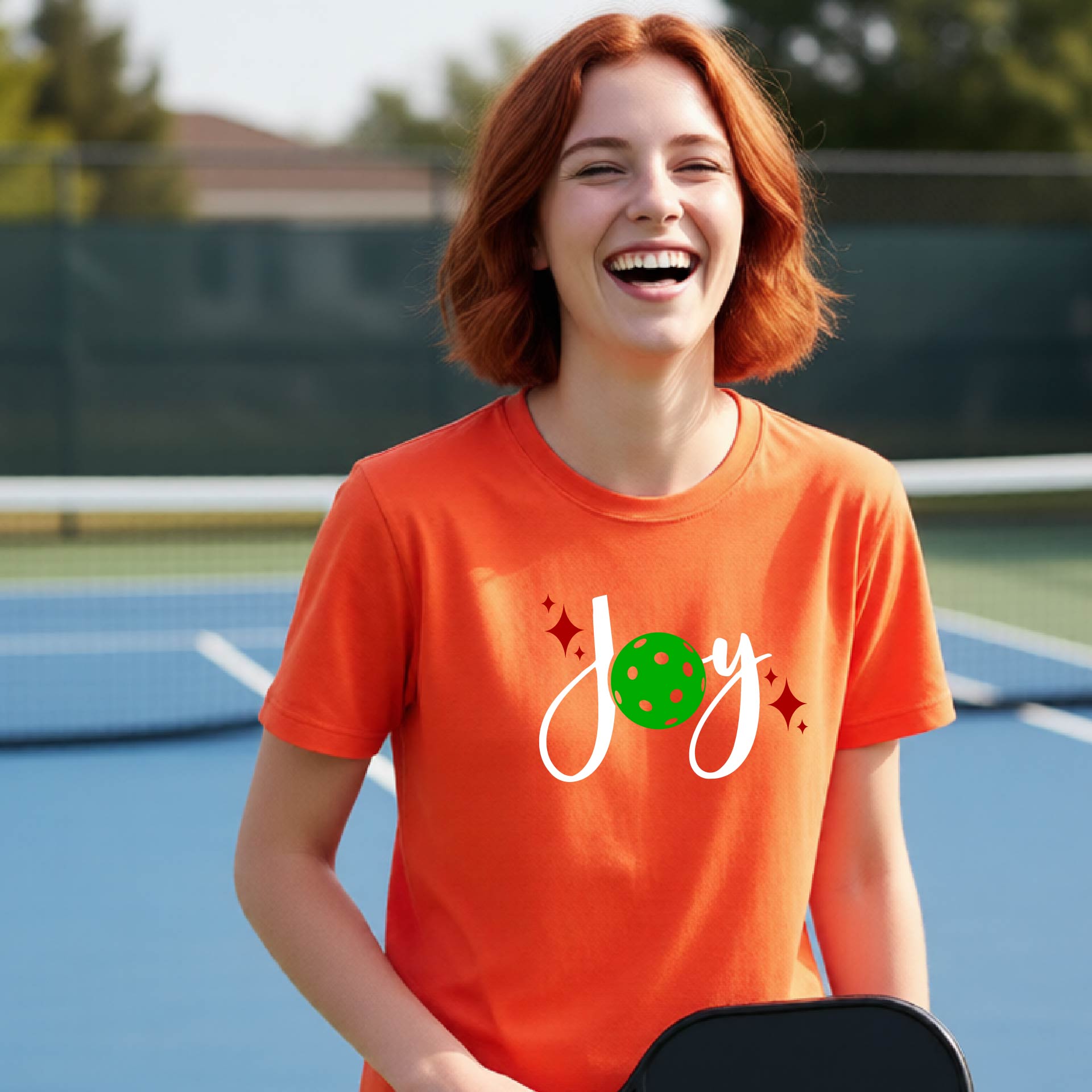 A young person laughing on an outdoor pickleball  court wearing an orange t-shirt with a white 'Joy' graphic and a green pickleball  motif.
