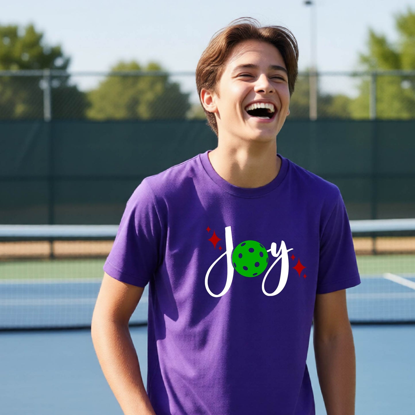 A young person laughing on an outdoor pickleball  court wearing a purple t-shirt with a white 'Joy' graphic and a green pickleball  motif.
