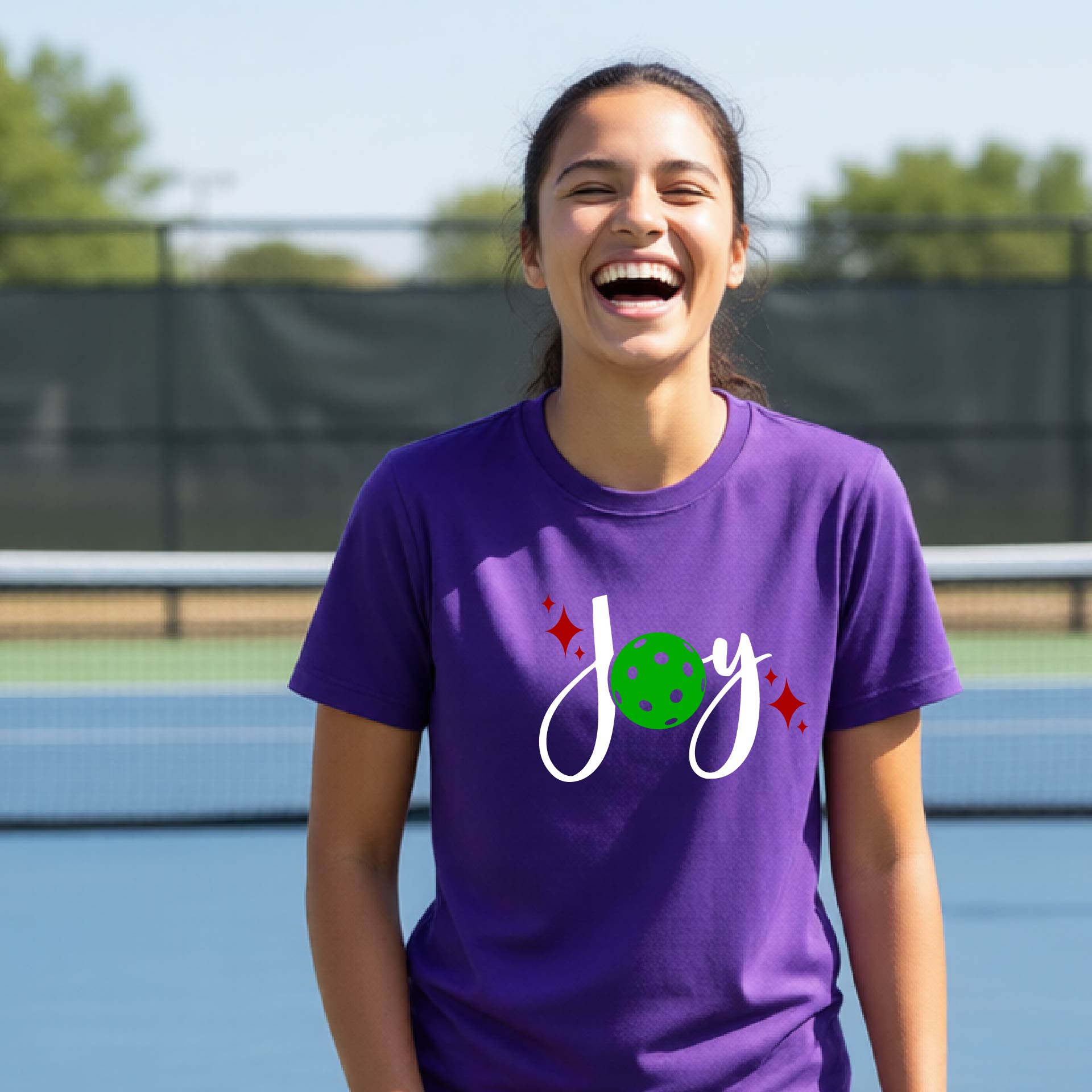A woman laughing on an outdoor pickleball  court wearing a purple t-shirt with a white 'Joy' graphic and a green pickleball  motif.
