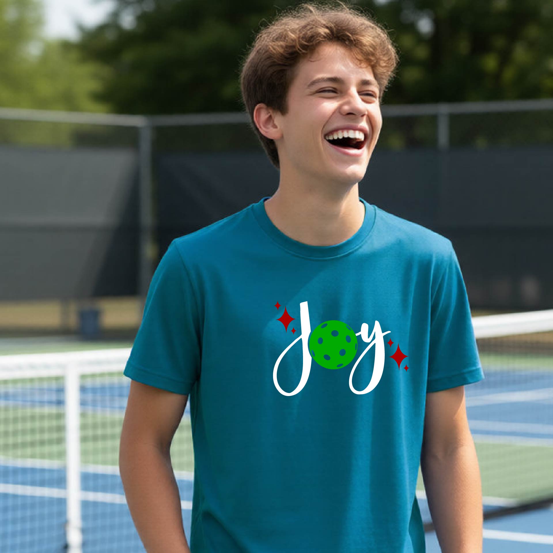 A young person laughing on an outdoor pickleball  court wearing a teal t-shirt with a white 'Joy' graphic and a green pickleball  motif.