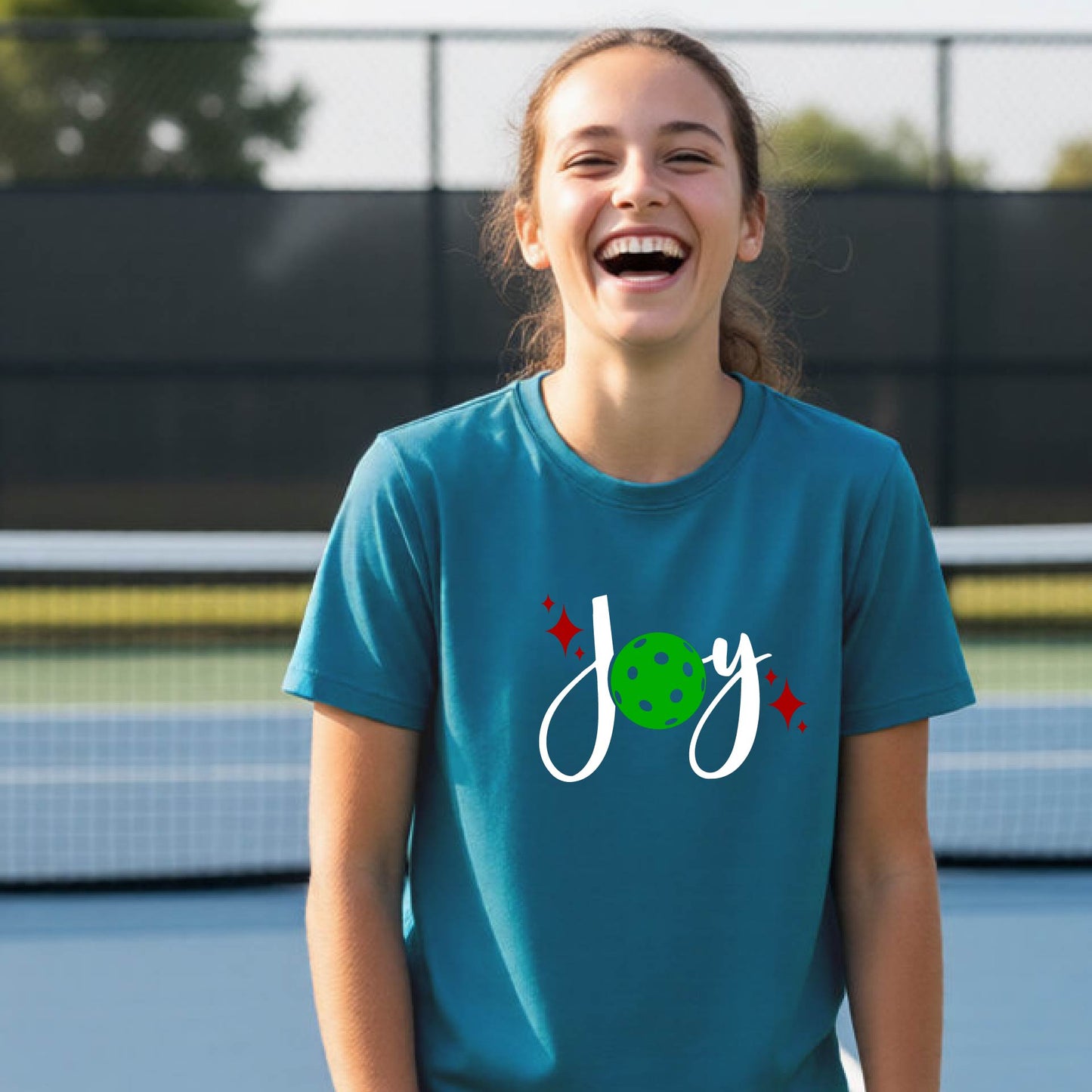 A young person laughing on an outdoor pickleball  court wearing a teal t-shirt with a white 'Joy' graphic and a green pickleball  motif.