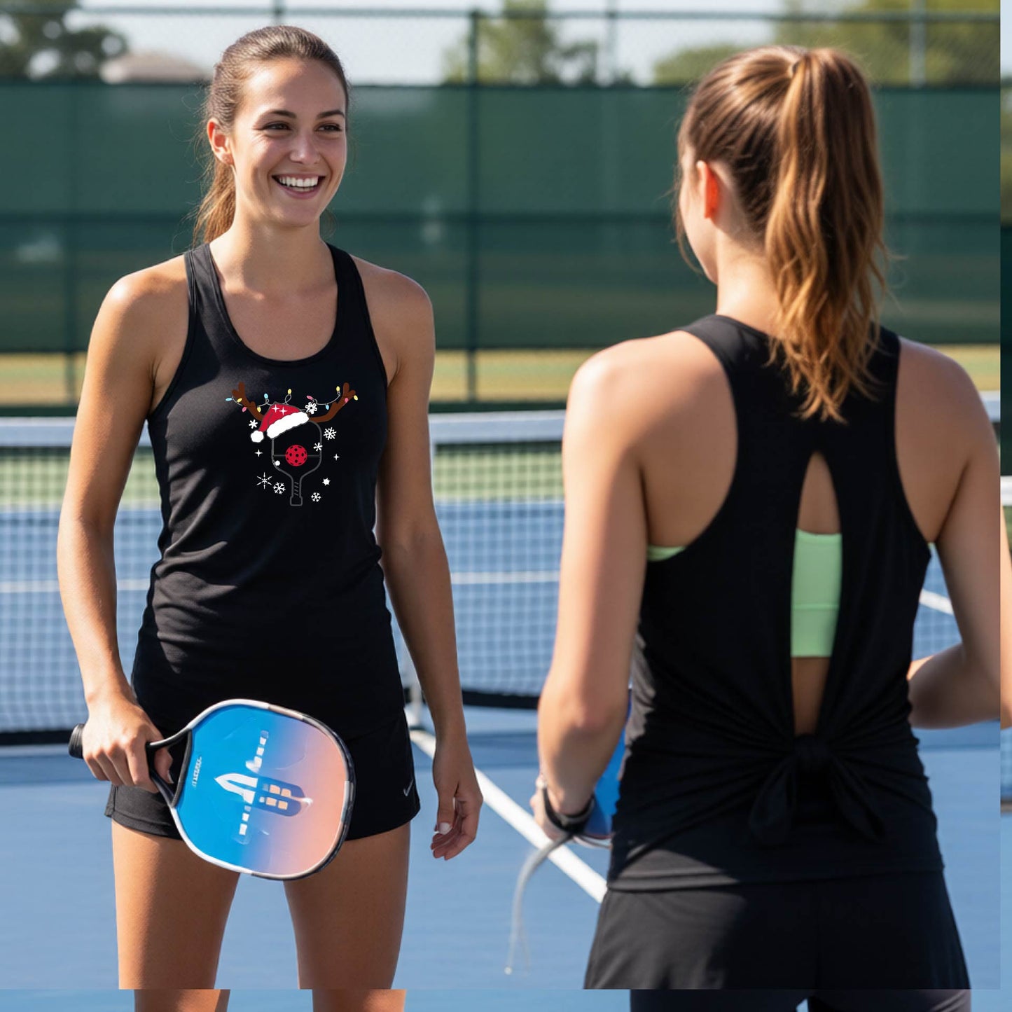 Two women on a blue pickleball court wearing matching black athletic tank tops with a cartoon pickleball reindeer paddle wearing a snata hat and antlers graphic. One shows the front of the top and the other shows the back with a tie-front detail.