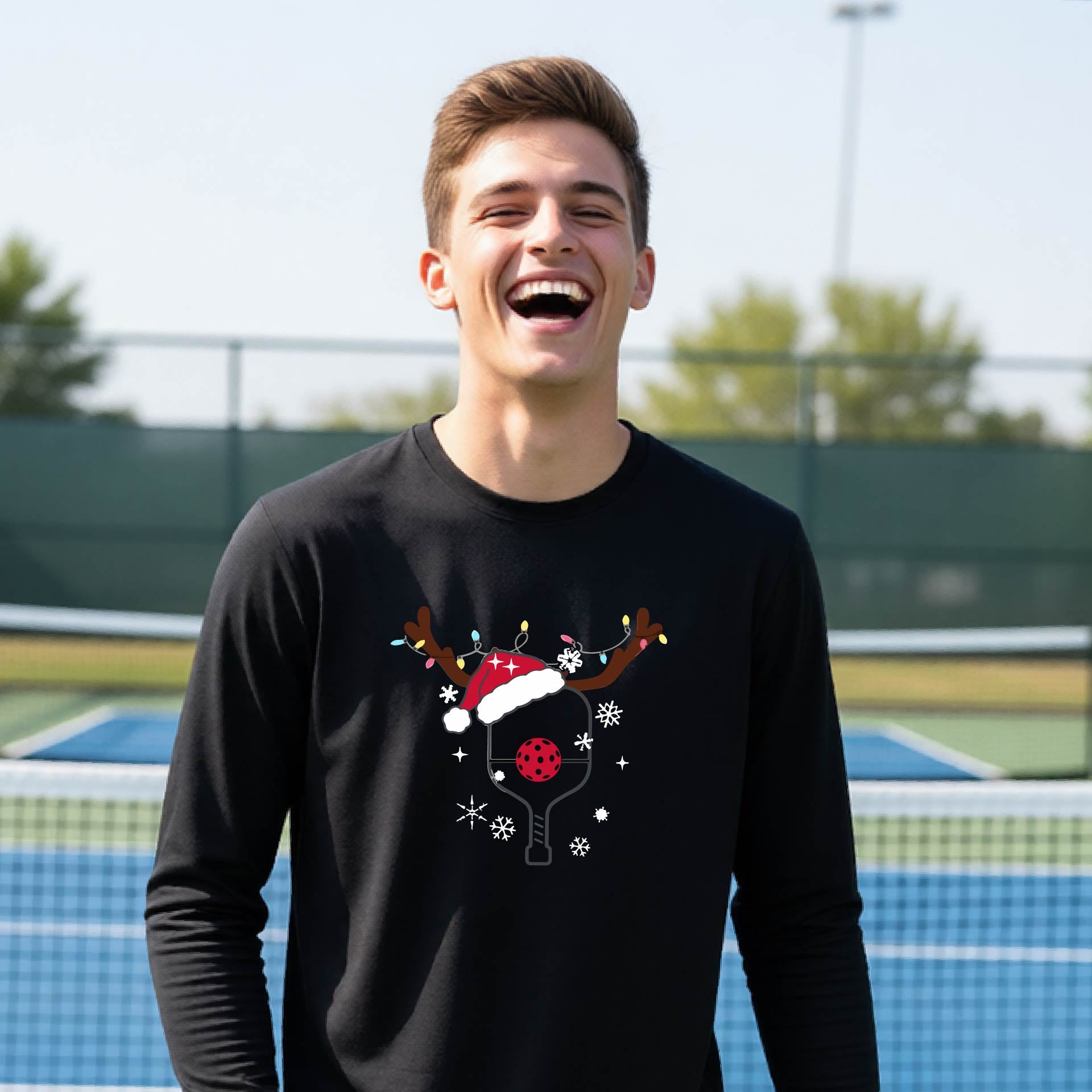 A young person laughing on an outdoor pickleball court, wearing a black long-sleeve shirt with a festive Christmas-themed picklabell graphic featuring a reindeer antler and Santa hat.