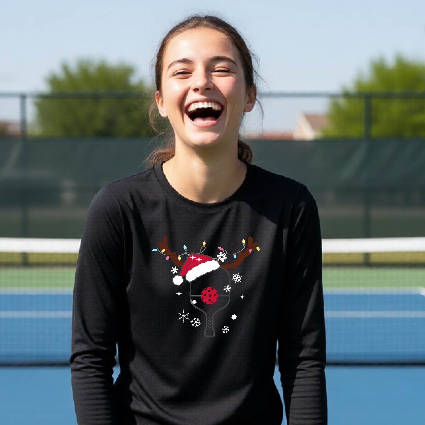 A young person laughing on an outdoor pickleball court, wearing a black long-sleeve shirt with a festive Christmas-themed picklabell graphic featuring a reindeer antler and Santa hat.