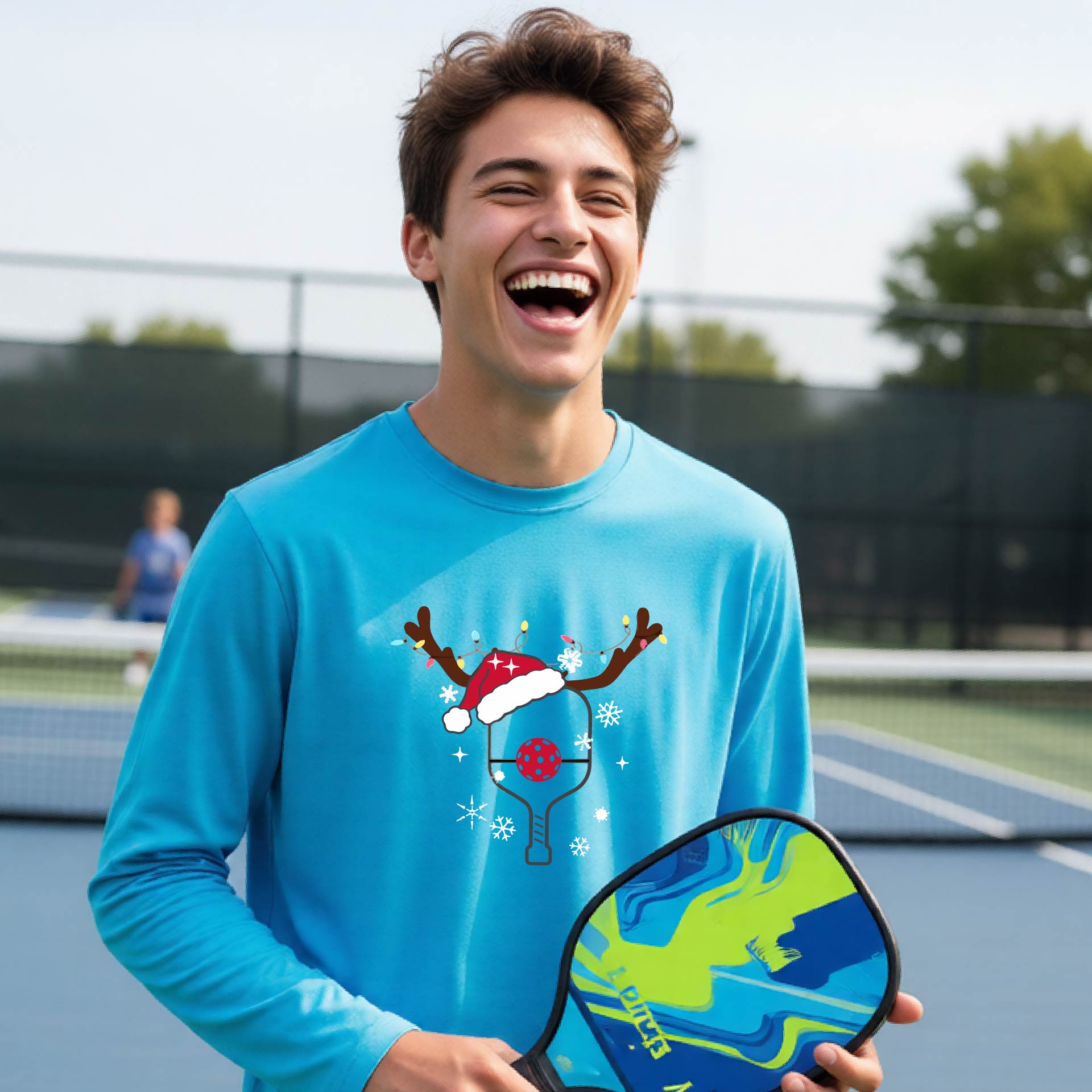 A young person laughing on an outdoor pickleball court, wearing a bright blue long-sleeve shirt with a festive Christmas-themed picklabell graphic featuring a reindeer antler and Santa hat.