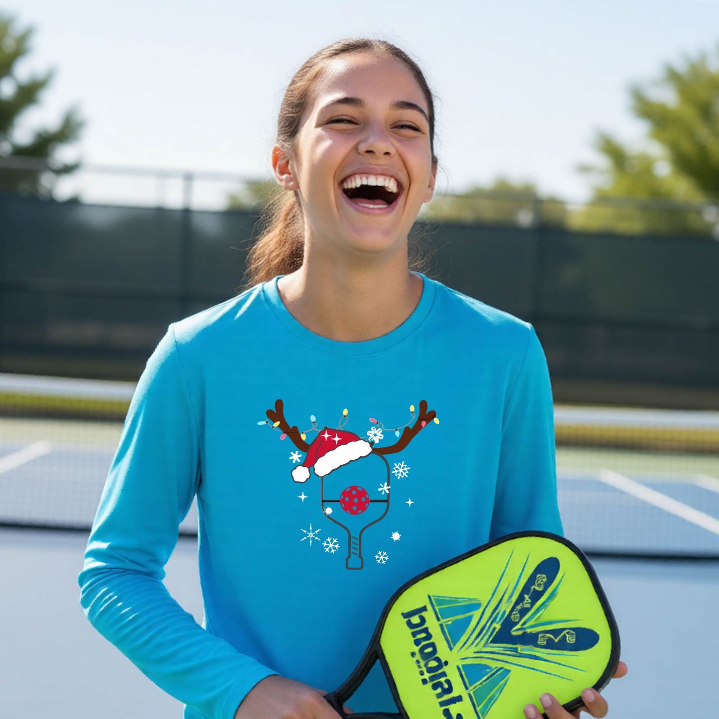 A young person laughing on an outdoor pickleball court, wearing a bright blue long-sleeve shirt with a festive Christmas-themed picklabell graphic featuring a reindeer antler and Santa hat.