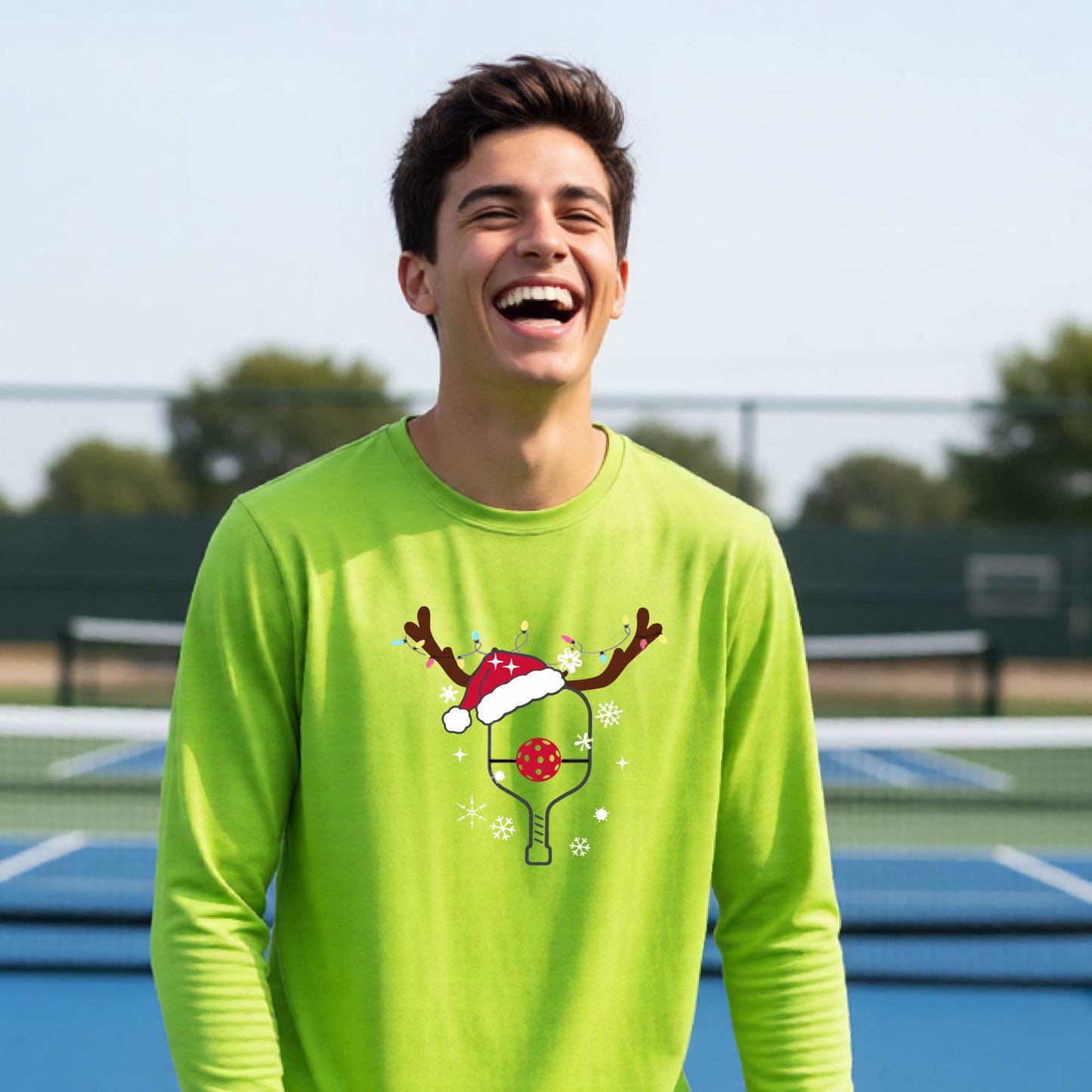 A young person laughing on an outdoor pickleball court, wearing a bright lime green long-sleeve shirt with a festive Christmas-themed picklabell graphic featuring a reindeer antler and Santa hat.
