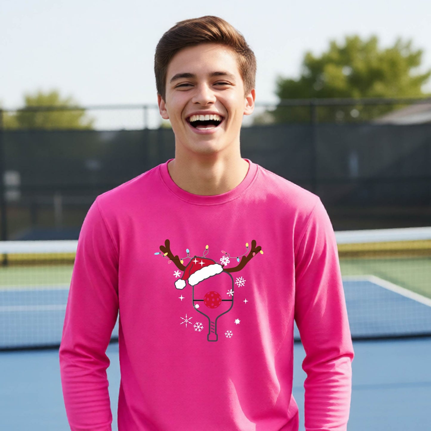 A young person laughing on an outdoor pickleball court, wearing a bright pink long-sleeve shirt with a festive Christmas-themed picklabell graphic featuring a reindeer antler and Santa hat.