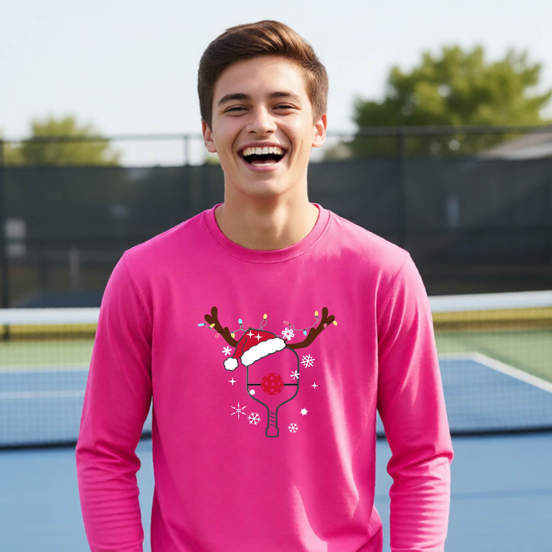 A young person laughing on an outdoor pickleball court, wearing a bright pink long-sleeve shirt with a festive Christmas-themed picklabell graphic featuring a reindeer antler and Santa hat.