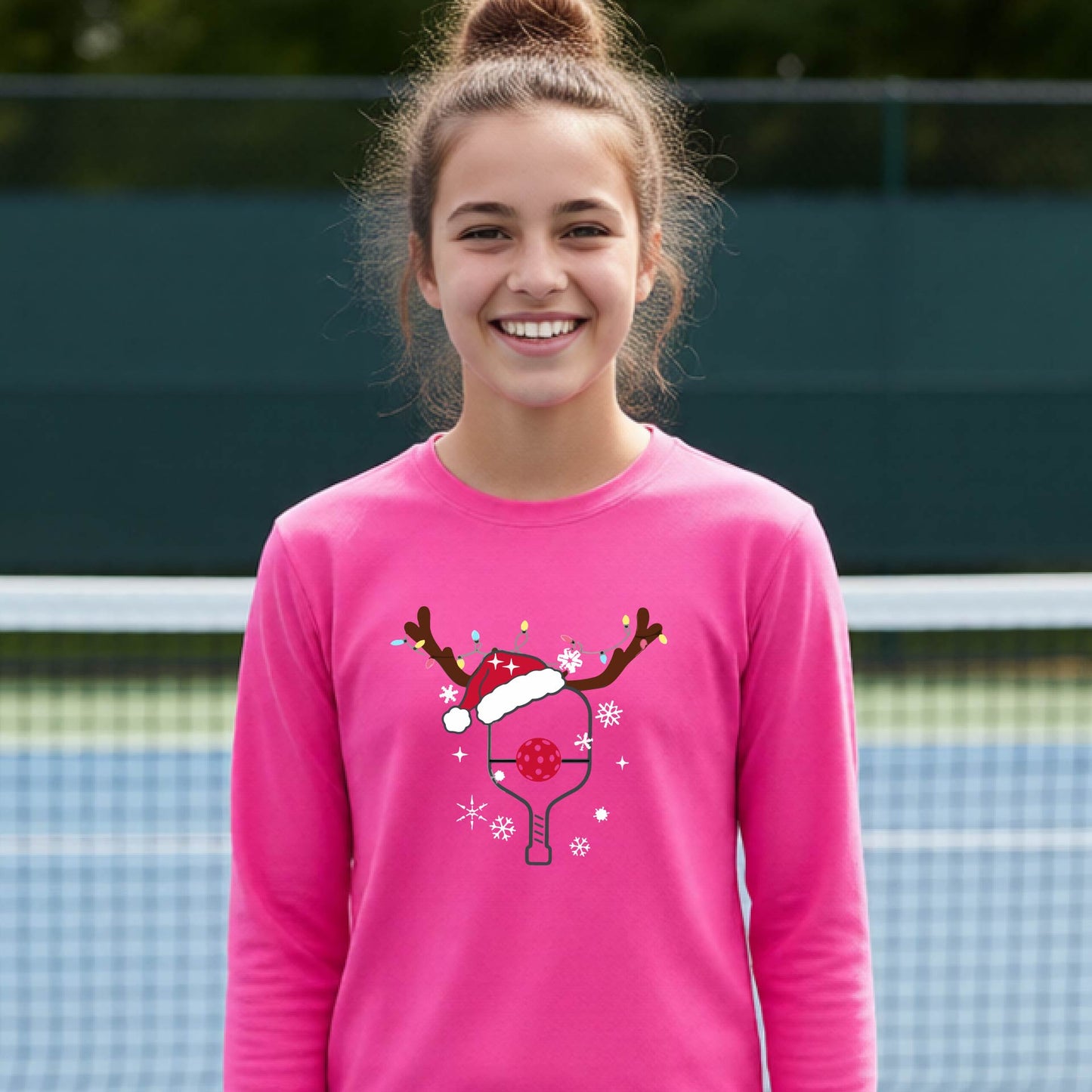 A young person laughing on an outdoor pickleball court, wearing a bright pink long-sleeve shirt with a festive Christmas-themed picklabell graphic featuring a reindeer antler and Santa hat.