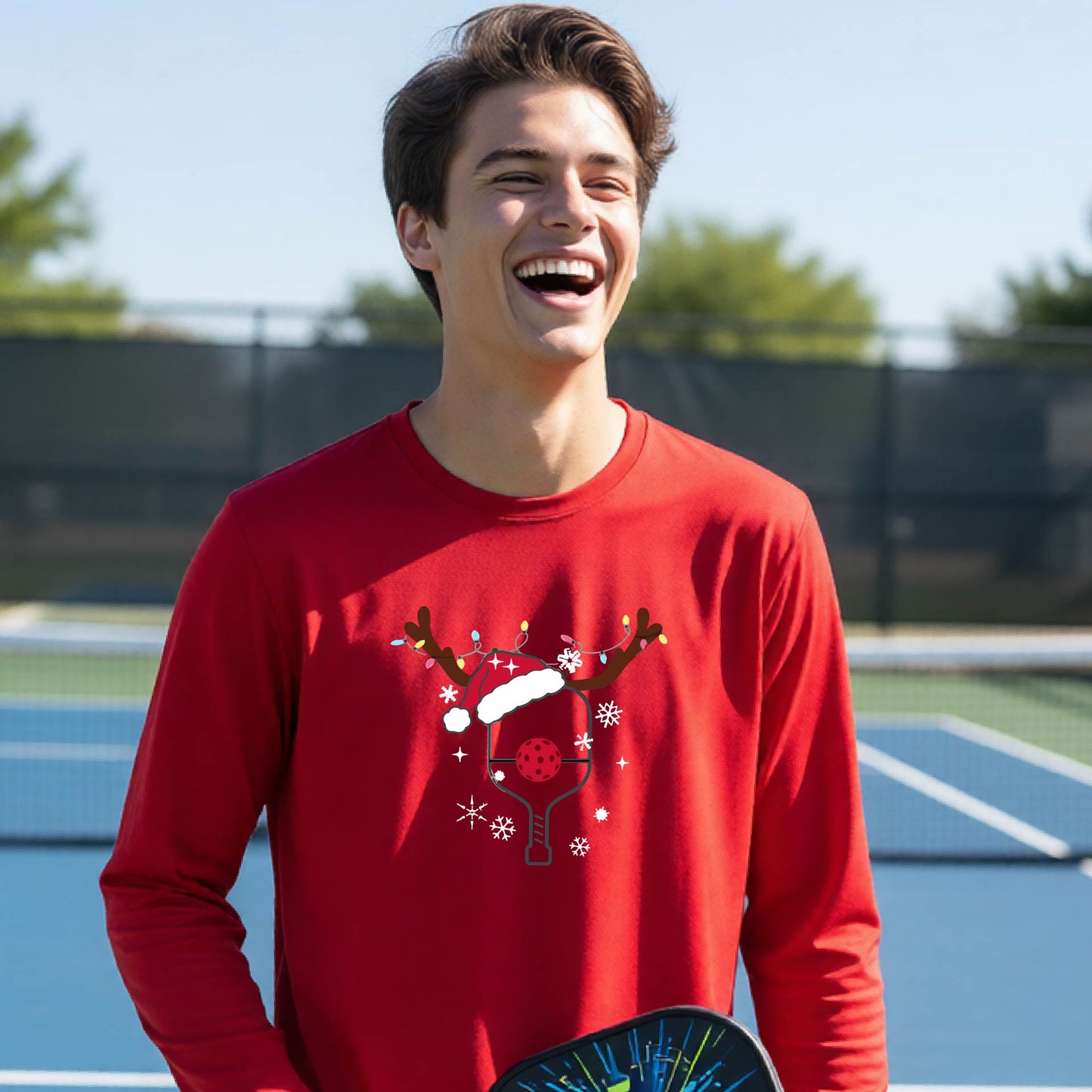 A young person laughing on an outdoor pickleball court, wearing a red long-sleeve shirt with a festive Christmas-themed picklabell graphic featuring a reindeer antler and Santa hat.