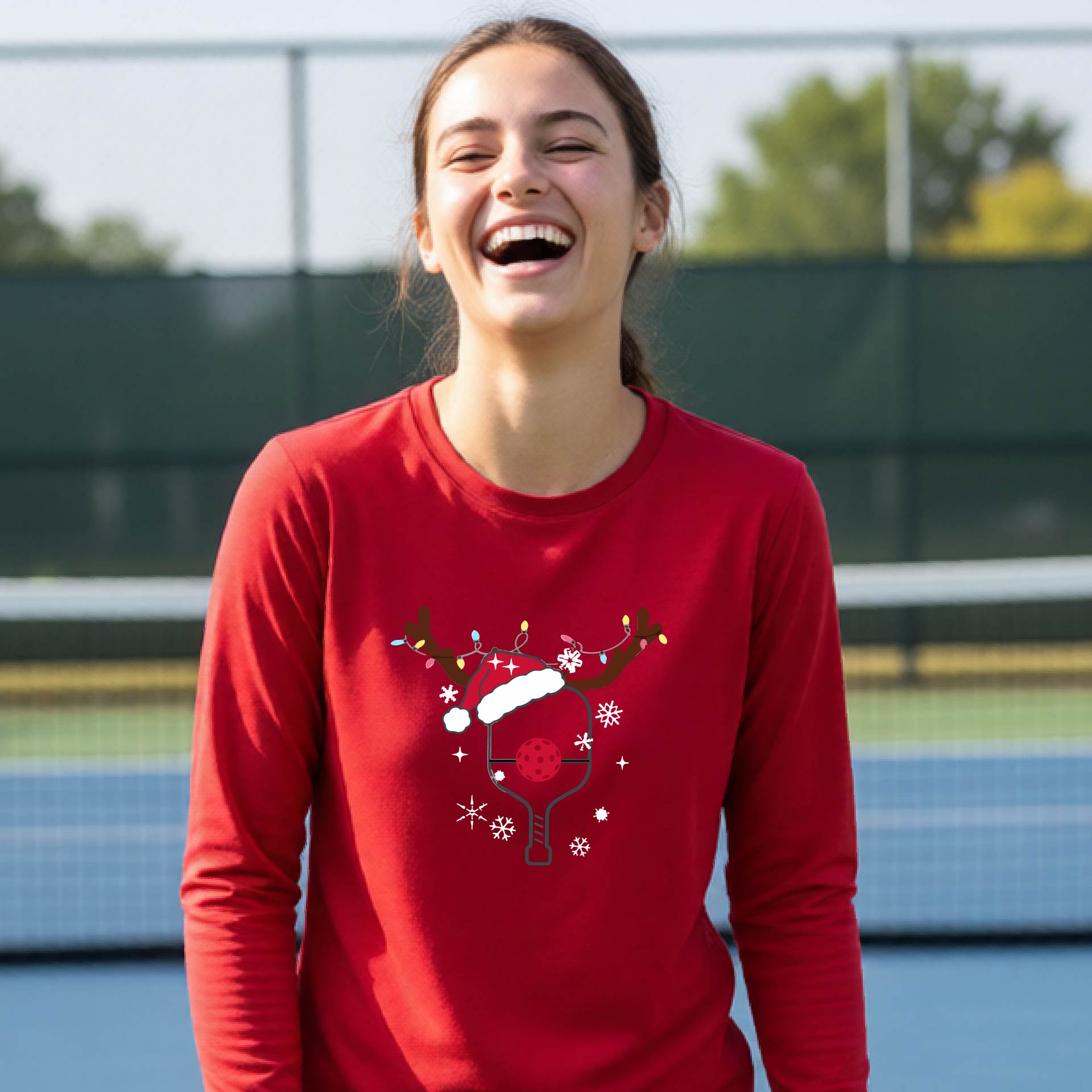 A young person laughing on an outdoor pickleball court, wearing a bright red long-sleeve shirt with a festive Christmas-themed picklabell graphic featuring a reindeer antler and Santa hat.