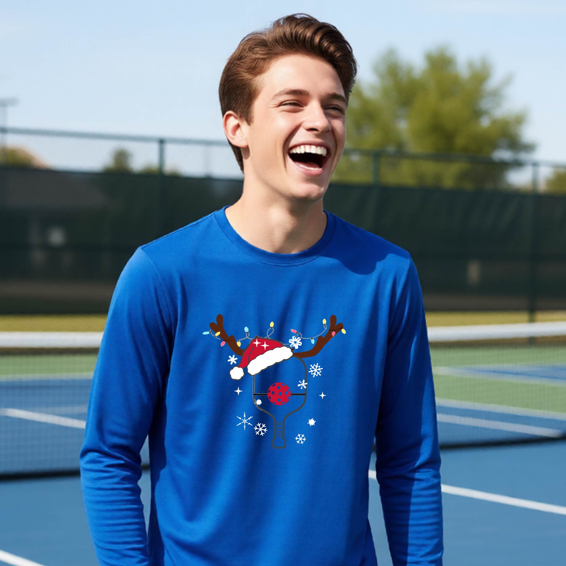 A young person laughing on an outdoor pickleball court, wearing a bright royal blue long-sleeve shirt with a festive Christmas-themed picklabell graphic featuring a reindeer antler and Santa hat.