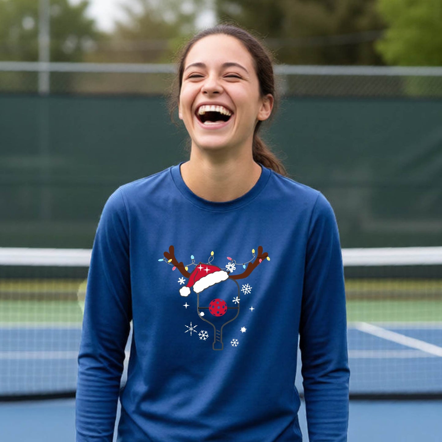 A young person laughing on an outdoor pickleball court, wearing a bright royal  blue long-sleeve shirt with a festive Christmas-themed picklabell graphic featuring a reindeer antler and Santa hat.