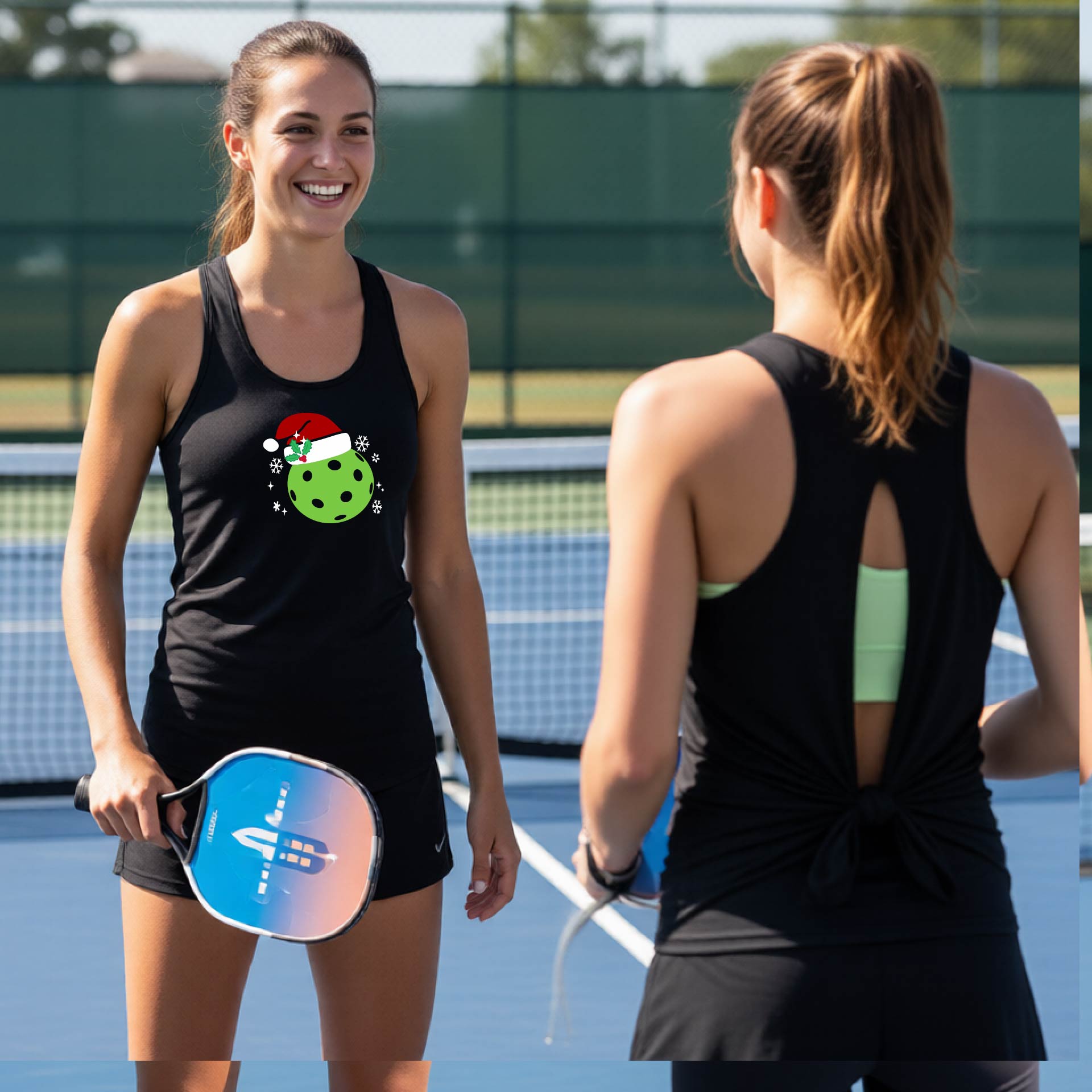 Two women on a pickleball court wearing matching black tank tops with different back designs, one with a green pickleball graphic wearing a santa hat and the other with a back tie.