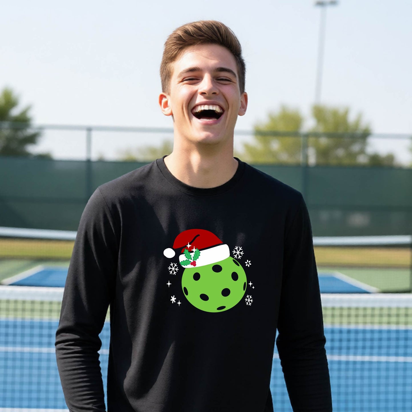 A young person laughing on an outdoor pickleball court wearing a black long-sleeve shirt with a green polka-dot pickleball graphic wearing a red Santa hat.