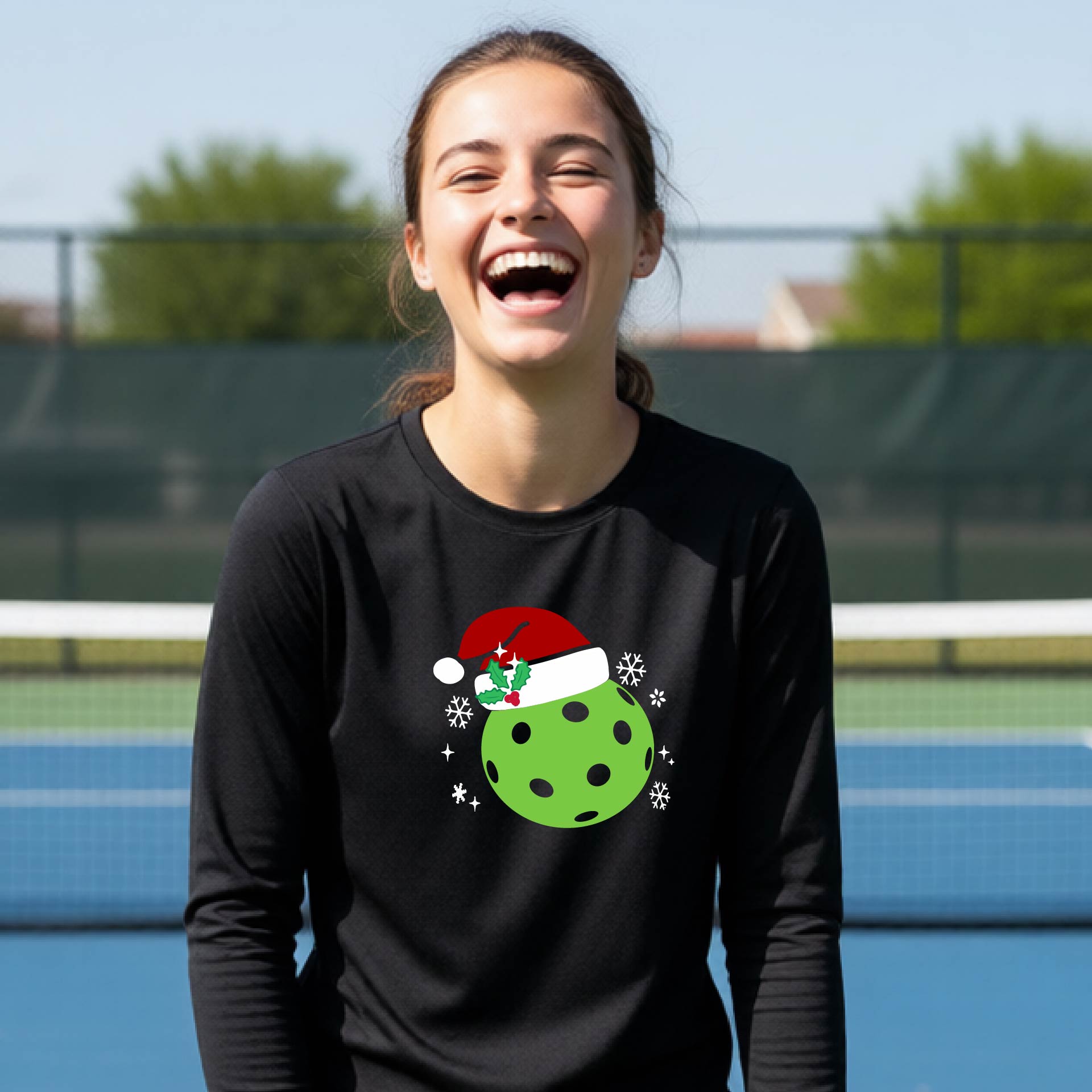 A young person laughing on an outdoor pickleball court wearing a black long-sleeve shirt with a green polka-dot pickleball graphic wearing a red Santa hat.