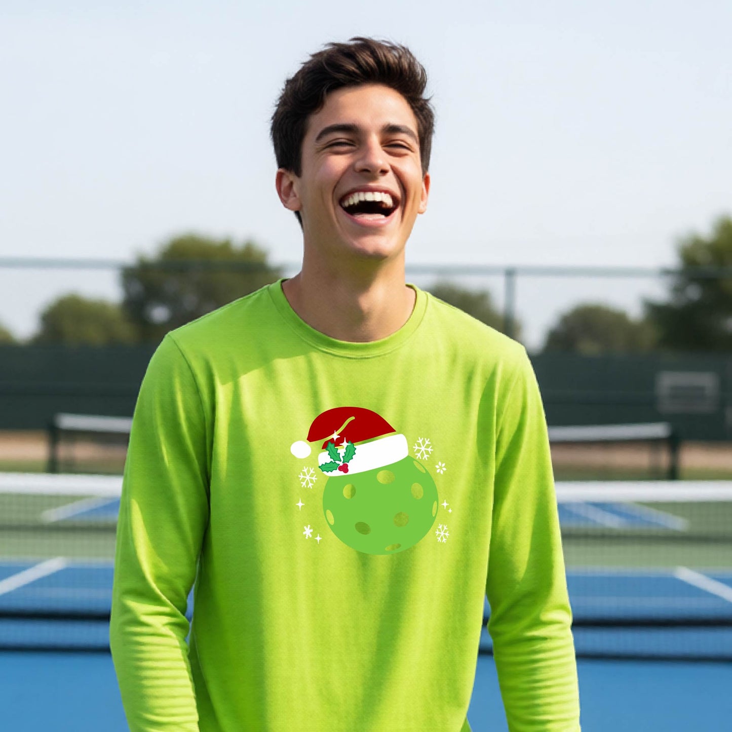 A young person laughing on an outdoor pickleball court wearing a lime green long-sleeve shirt with a green polka-dot pickleball graphic wearing a red Santa hat.