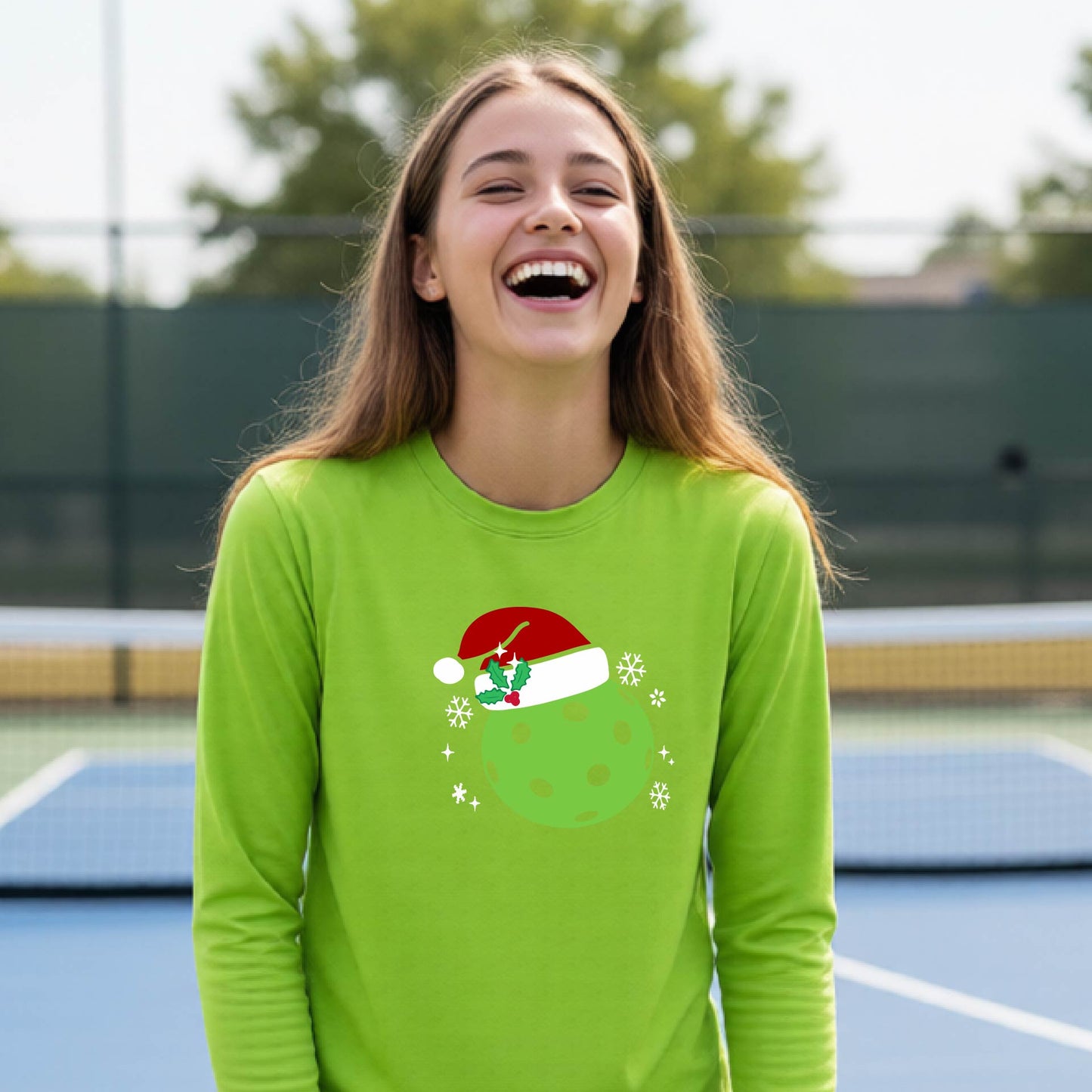 A young person laughing on an outdoor pickleball court wearing a lime green long-sleeve shirt with a green polka-dot pickleball graphic wearing a red Santa hat.